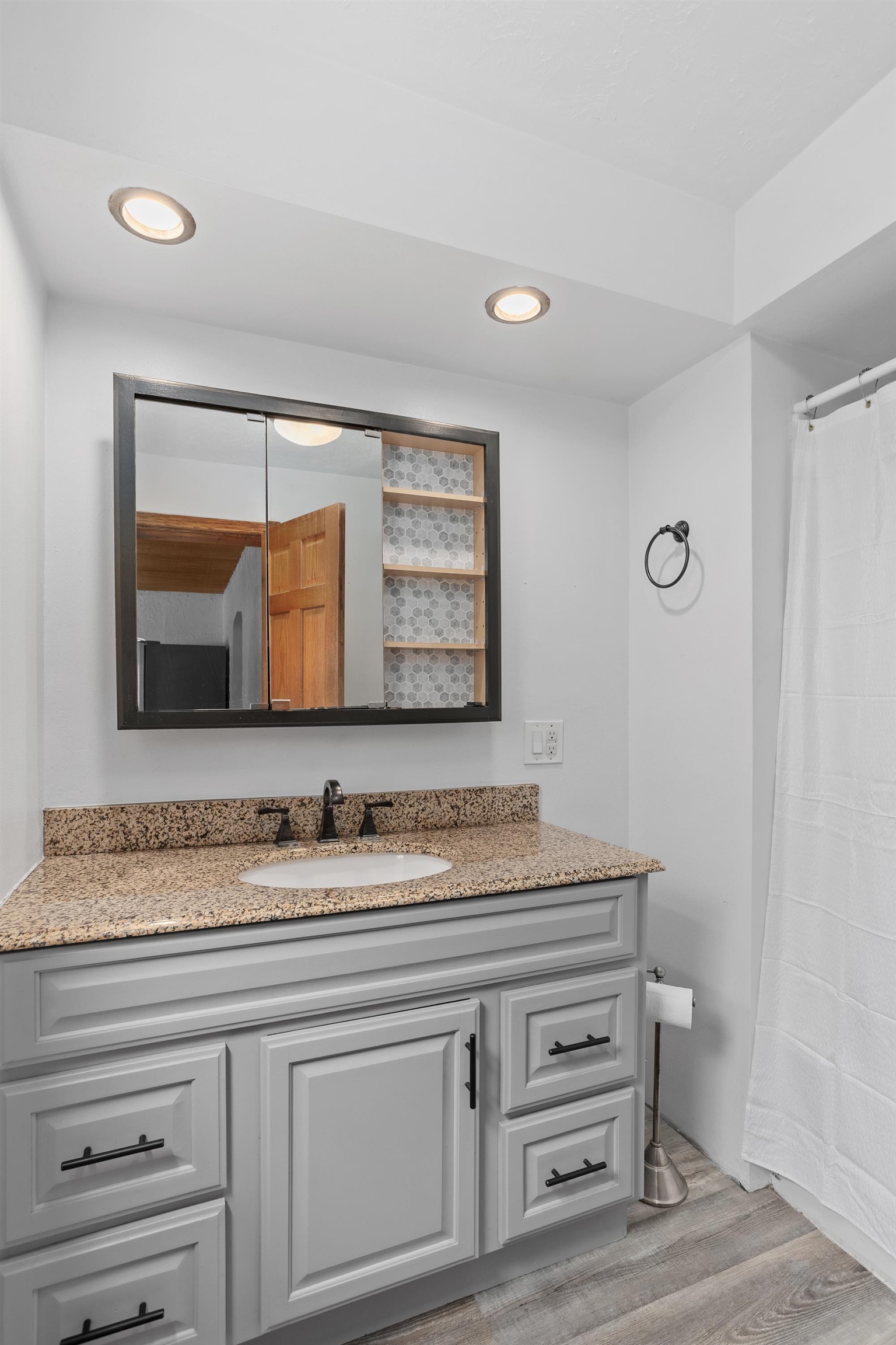 Bathroom vanity with a speckled granite counter, gray cabinetry, and a dark-framed mirror with tiled shelves.