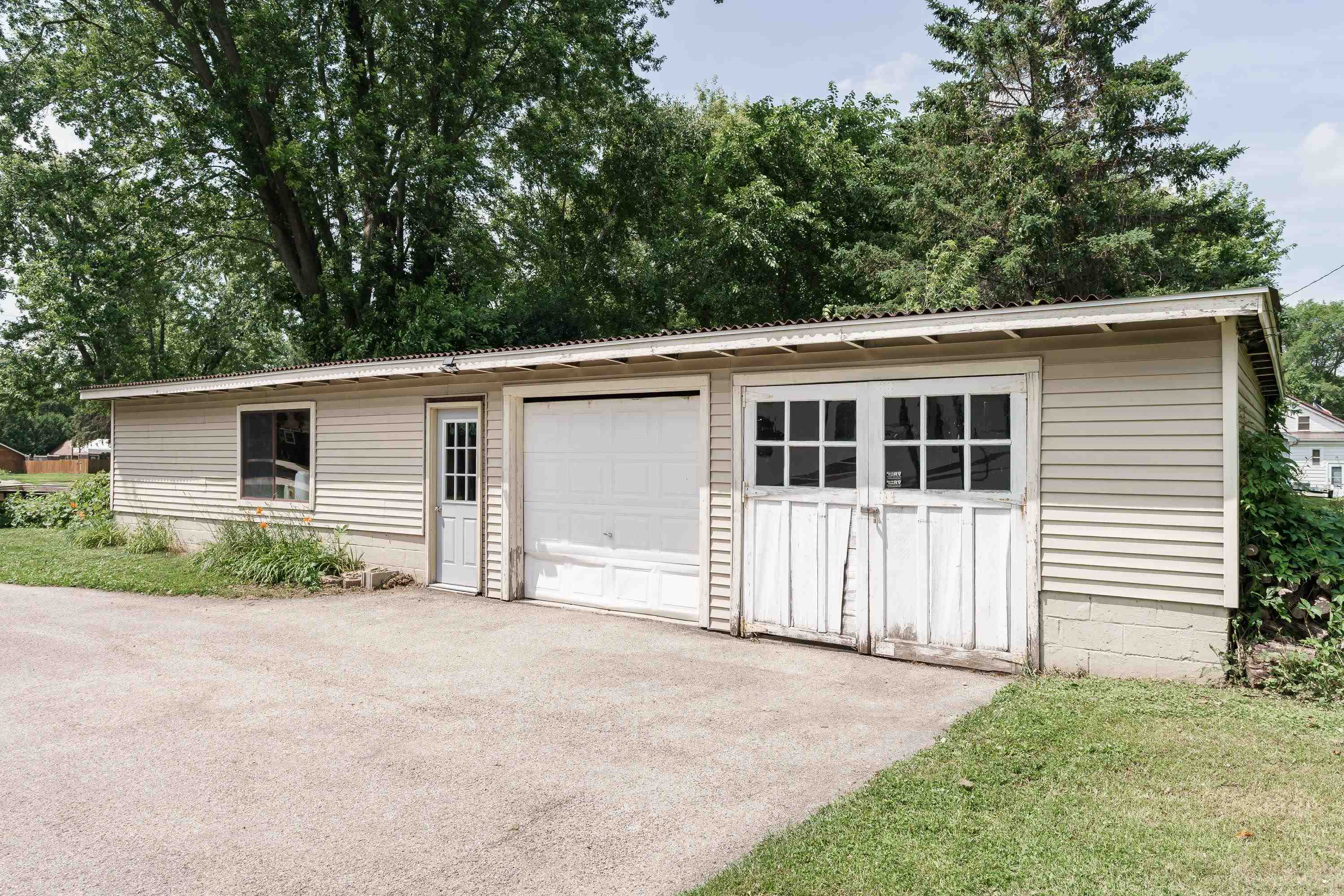 Detached beige garage with a white overhead door and weathered double doors beside a gravel driveway.