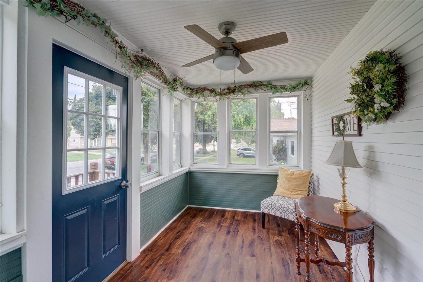Enclosed sunroom with a navy door, wraparound windows, green wainscoting, and dark wood floors.