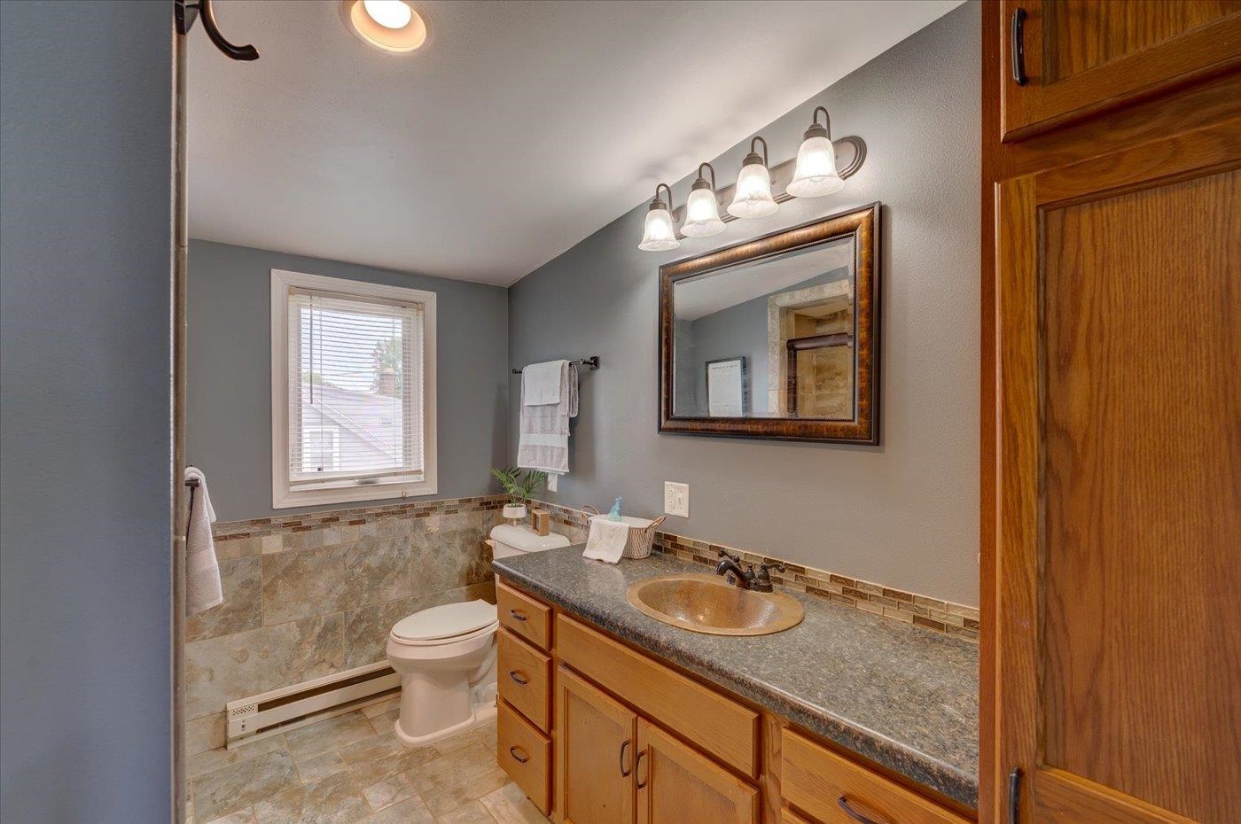 Bathroom with a wood vanity and oval sink, tiled walls, framed mirror, and window with blinds.