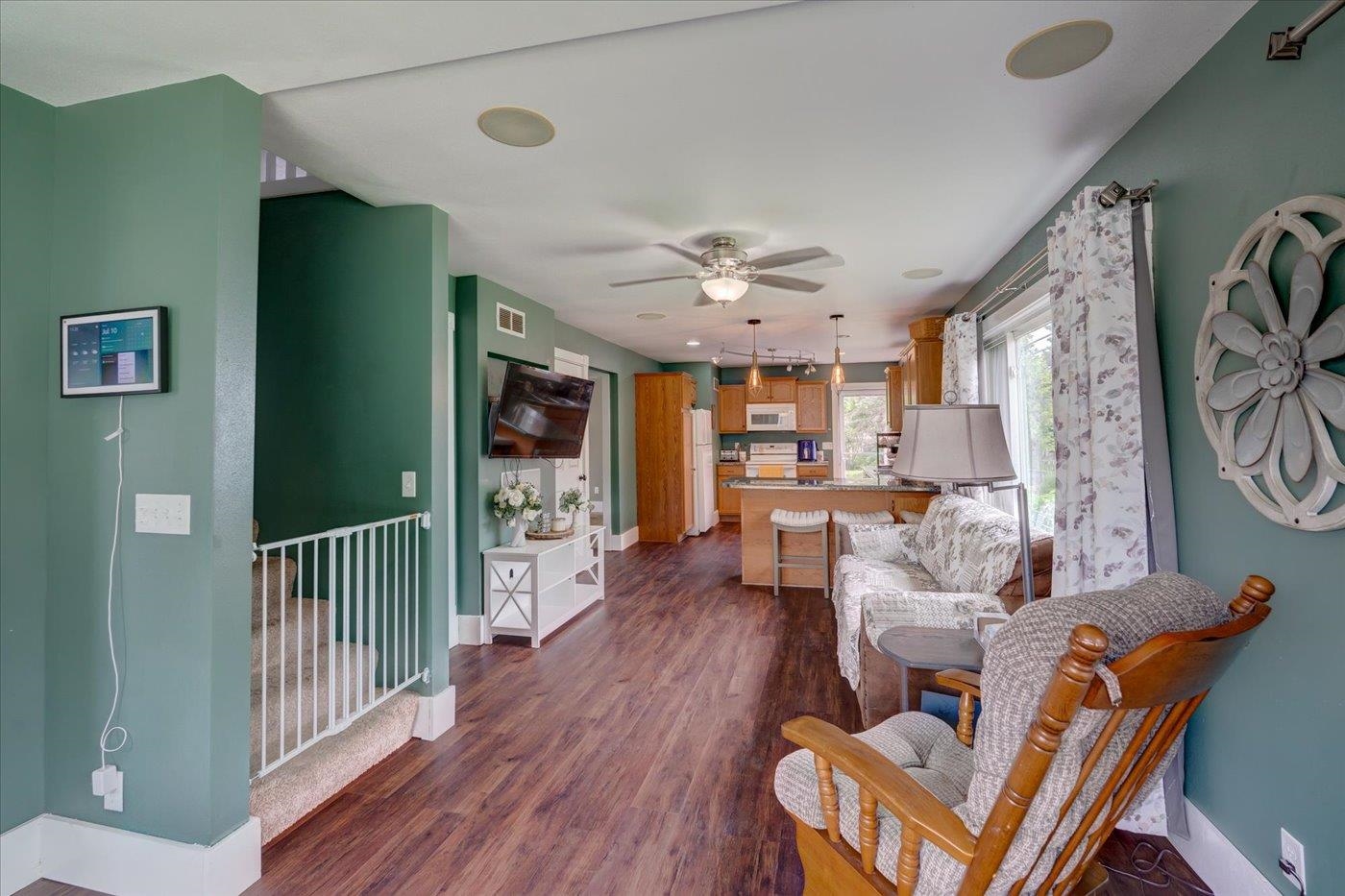 Open-plan living room and kitchen with teal walls, wood floors, and patterned seating by large windows.