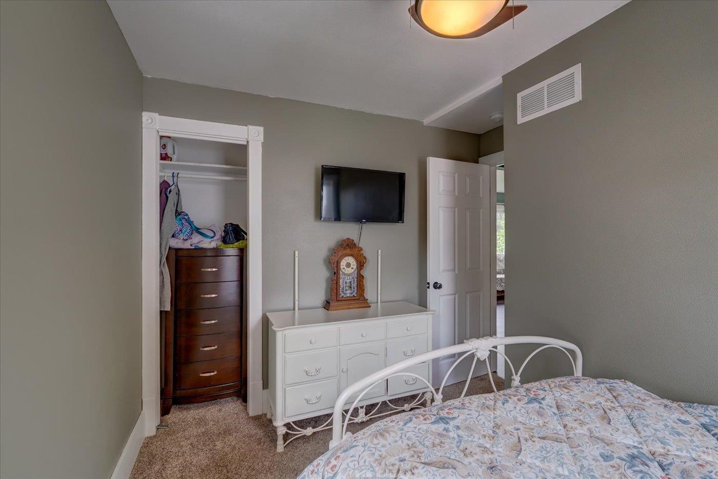 Bedroom with a white metal bed, floral quilt, dresser, wall-mounted TV, and an open closet with a dark wood chest.