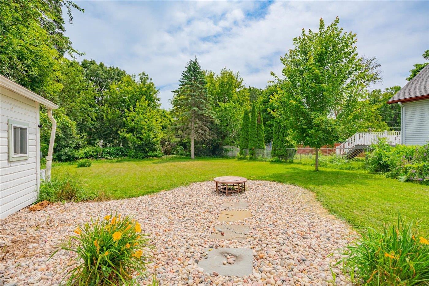 Backyard with a stone-covered patio, circular fire pit, broad lawn, and mature trees.
