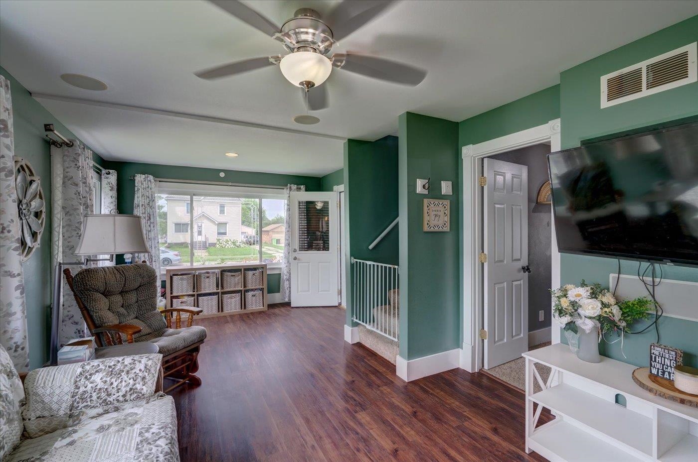 Living room with green walls, wood-look floors, a ceiling fan, seating, and a large window.