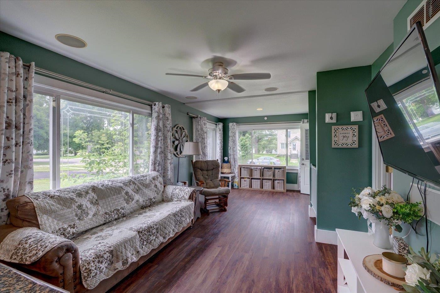 Living room with green walls, large curtained windows, a patterned sofa, and wood-look floors.