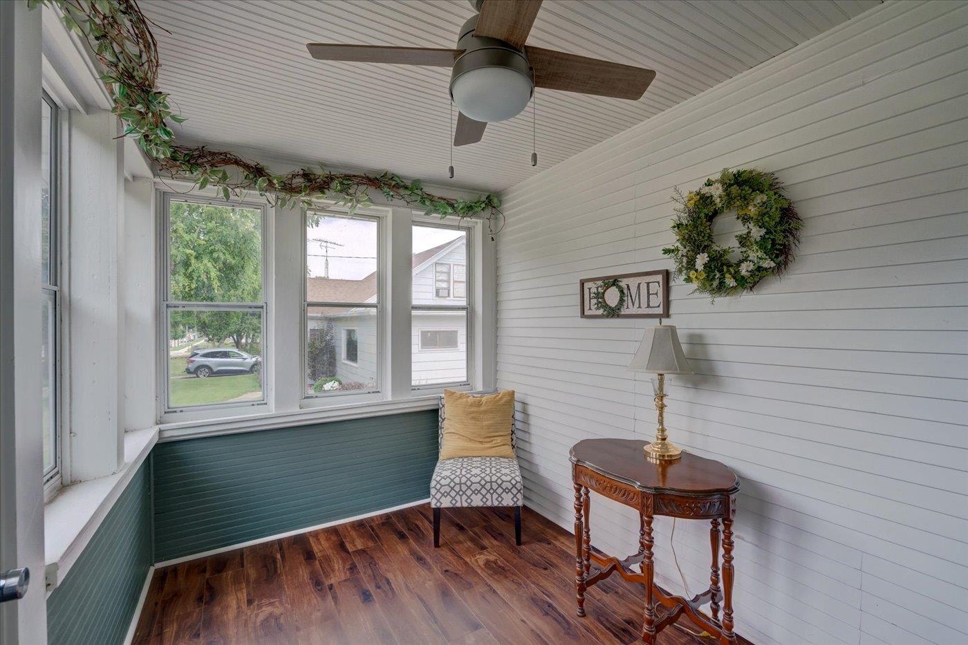 Enclosed porch with tall windows, a cushioned chair, ceiling fan, and wreath above a HOME sign.
