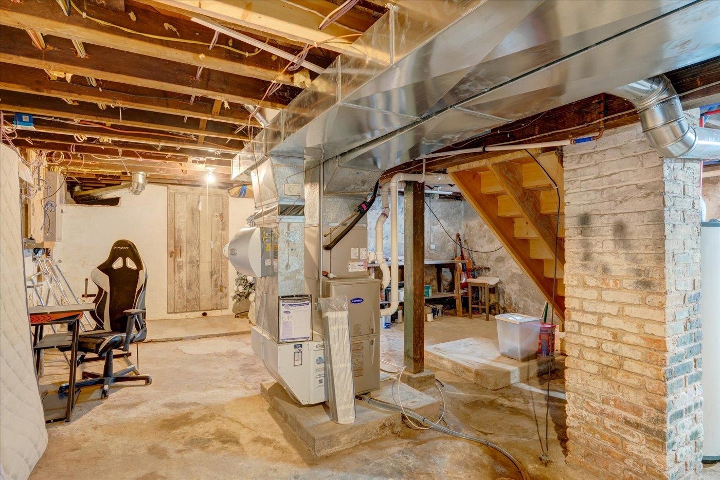 Unfinished basement with exposed ductwork, a central furnace, stone pillar, and staircase.