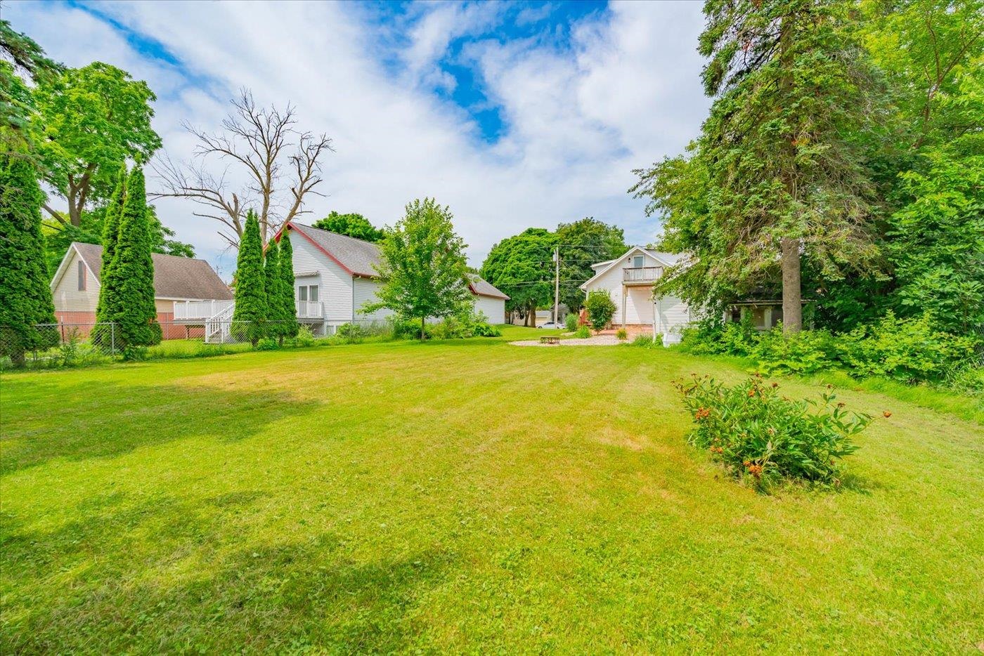 Spacious grassy backyard bordered by mature trees, with neighboring houses and a small gravel seating area.