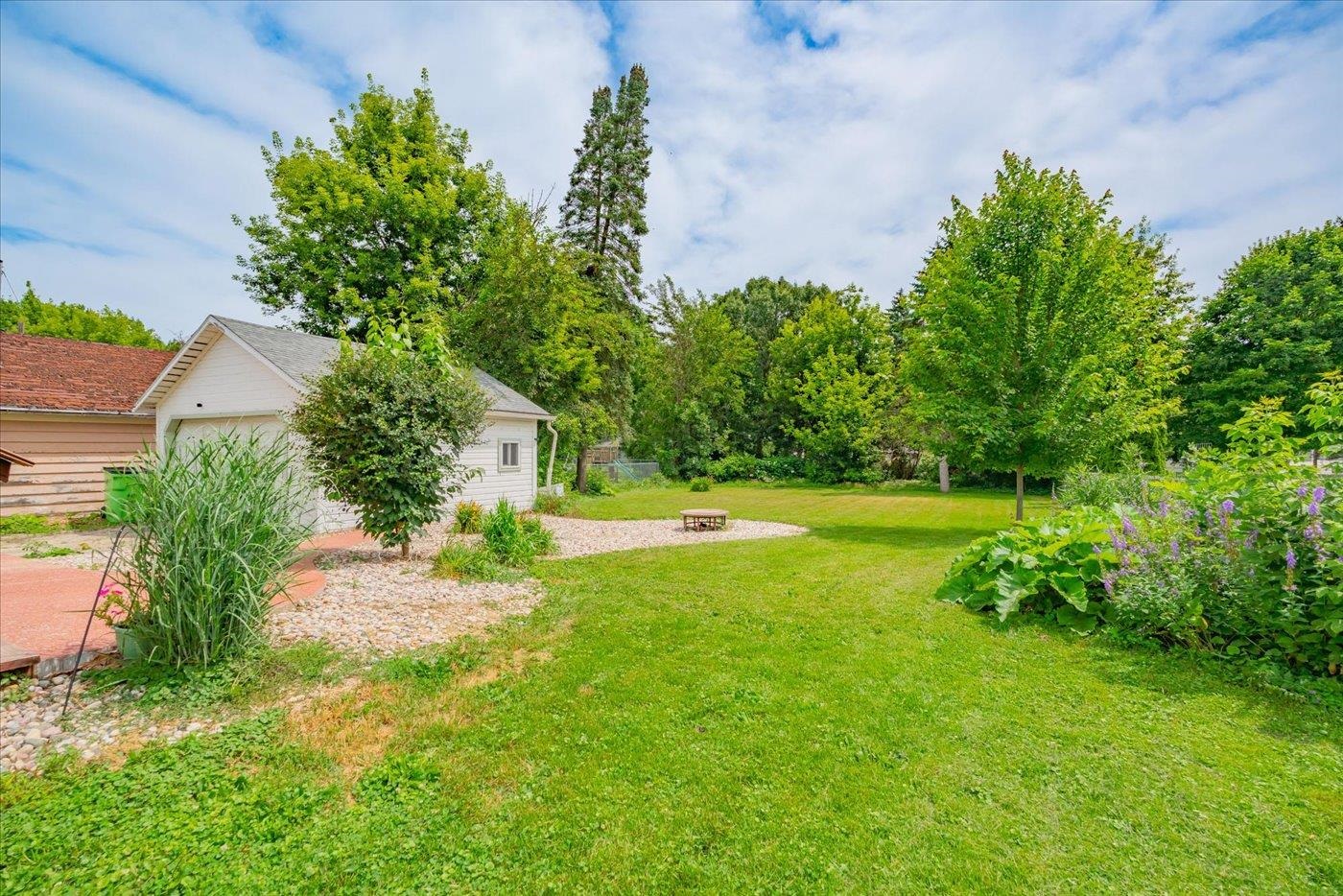 Landscaped backyard with a broad lawn, mature trees, stone beds, and a small patio.