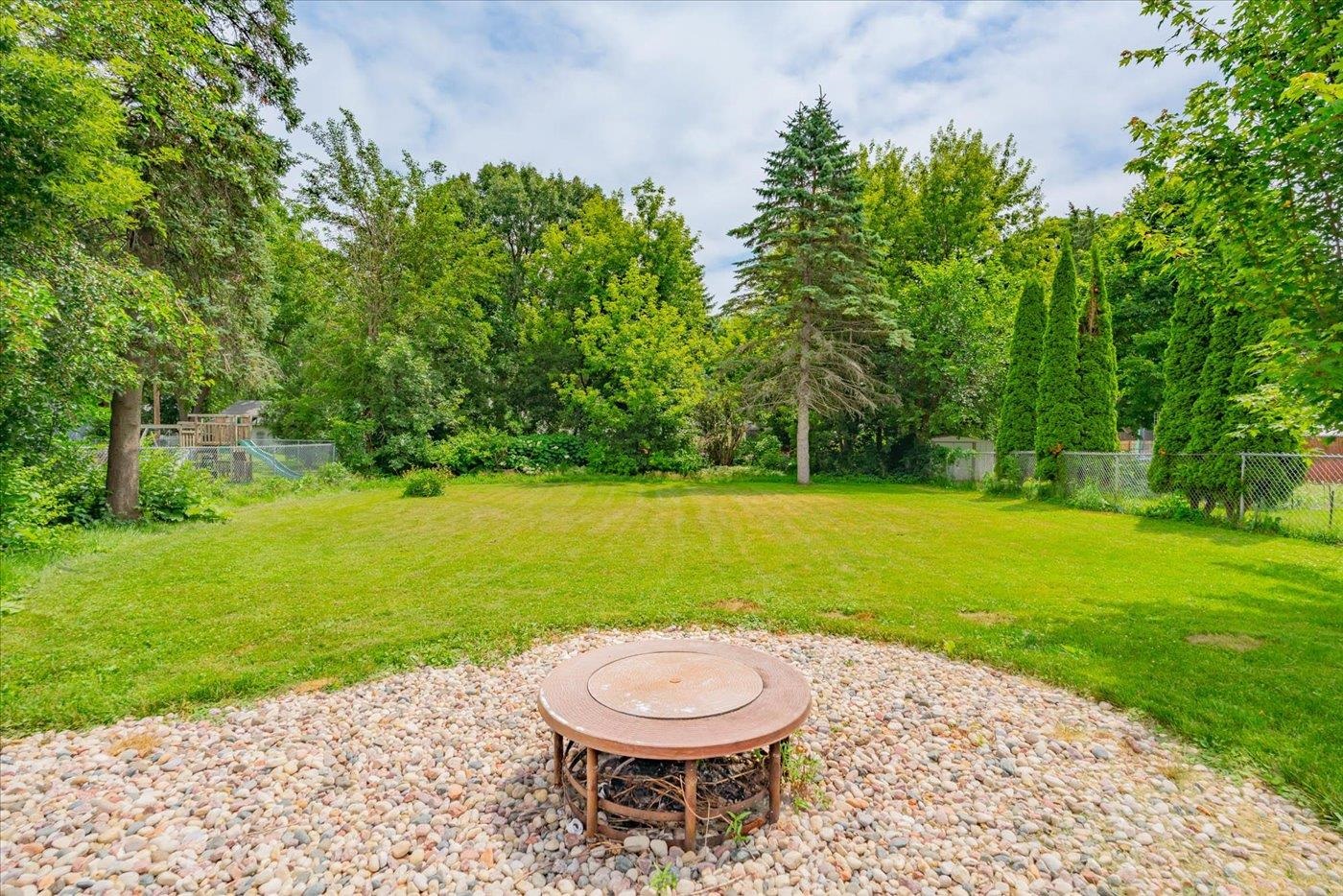 Backyard with a wide lawn, mature trees, and a round fire pit in a river-rock bed.