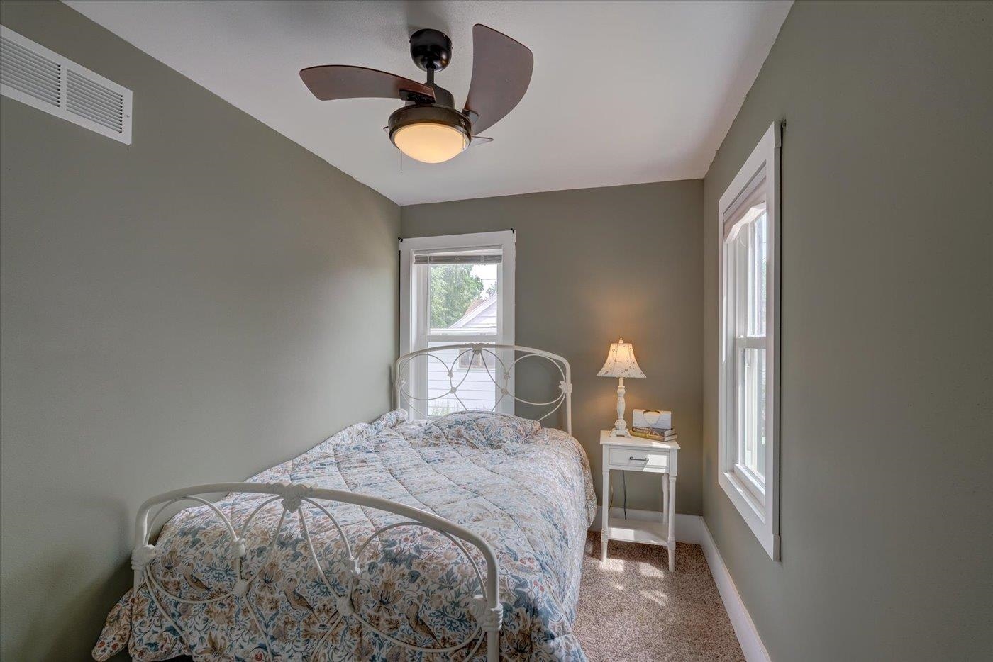 Bedroom with a white metal bed, floral quilt, two windows, and ceiling fan with light.