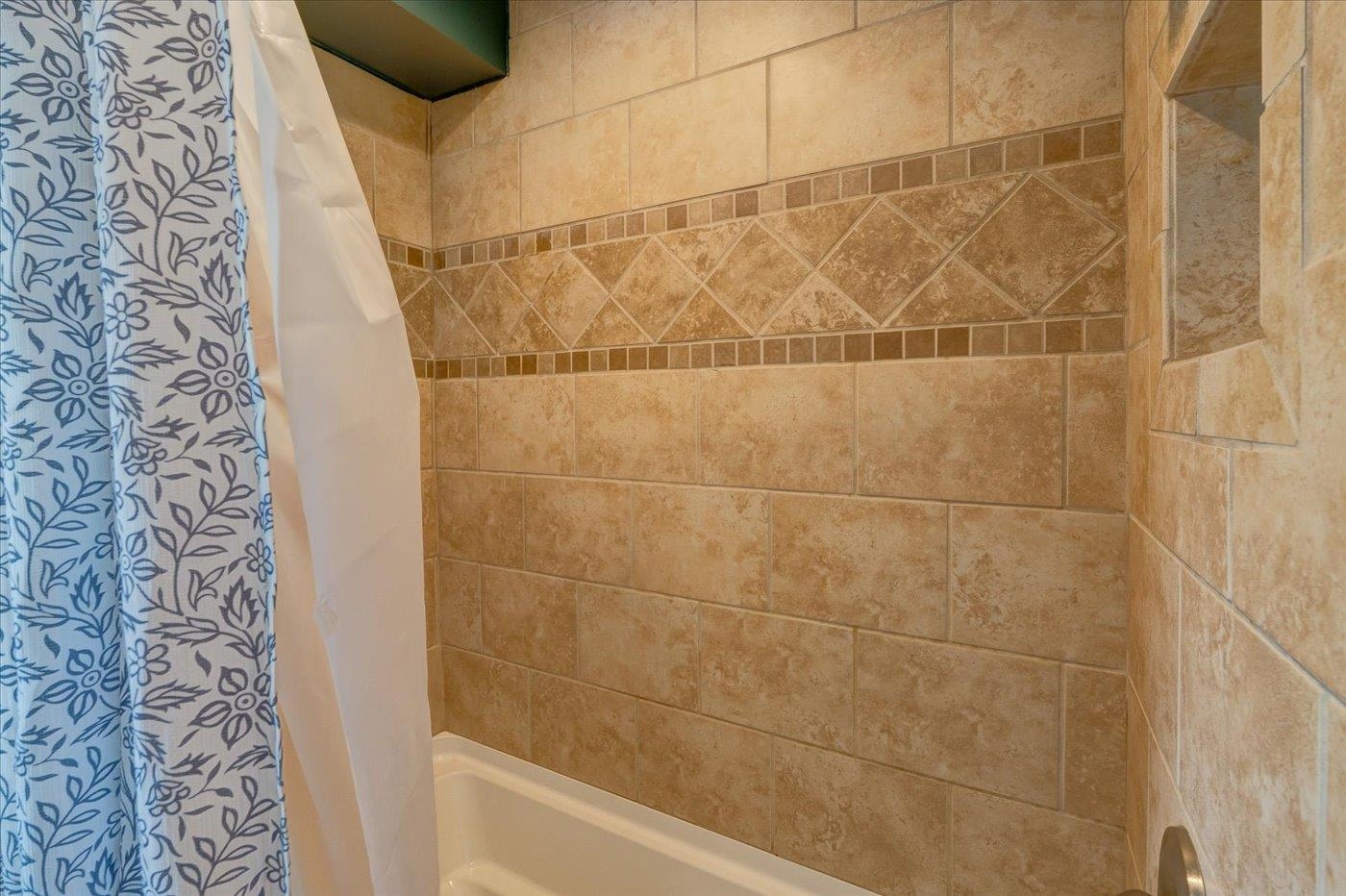 Bathtub with tan tiled surround and a diamond-pattern accent band beside blue floral shower curtains.