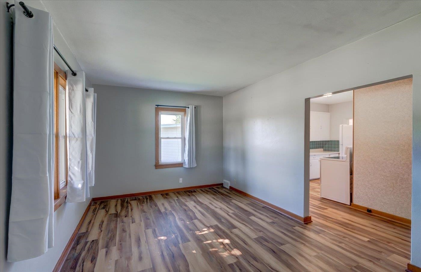 Empty living room with wood-look floors, curtained windows, and a doorway to the kitchen.