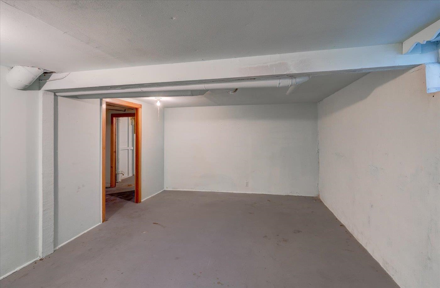Empty basement room with a low ceiling, exposed ductwork, and a wood-trimmed doorway.