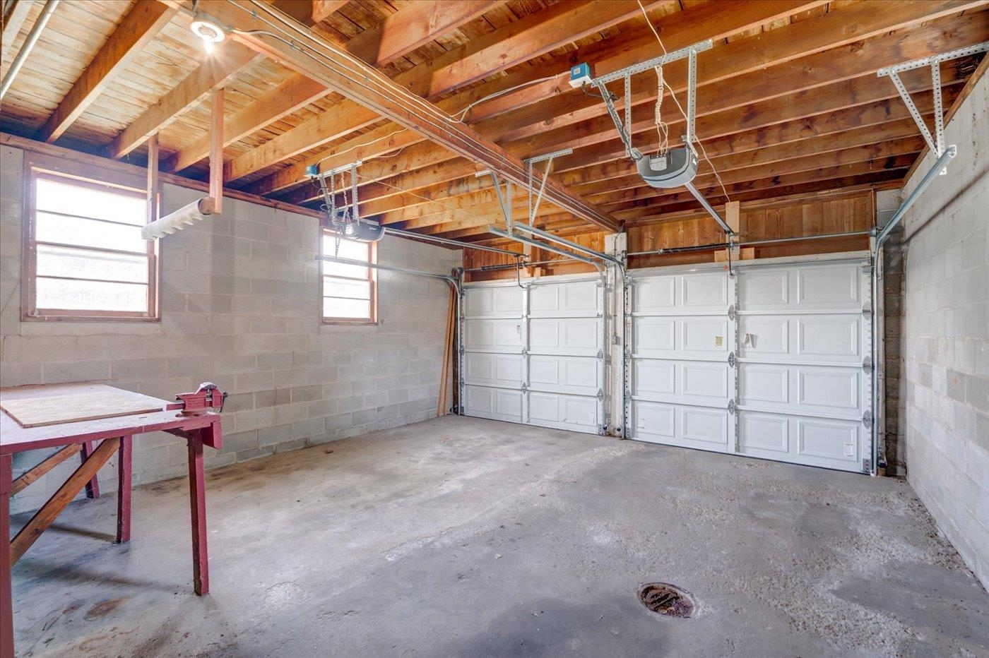 Garage with two white overhead doors, exposed wooden roof framing, and a concrete floor drain.