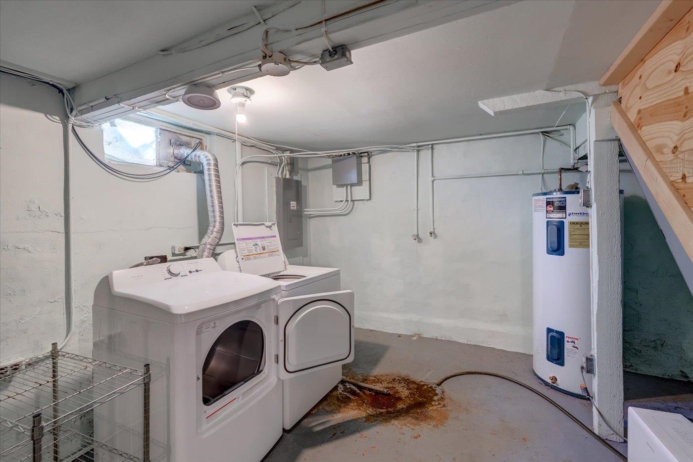 Basement laundry area with a washer and dryer, water heater, exposed pipes, and a rust-colored floor stain.