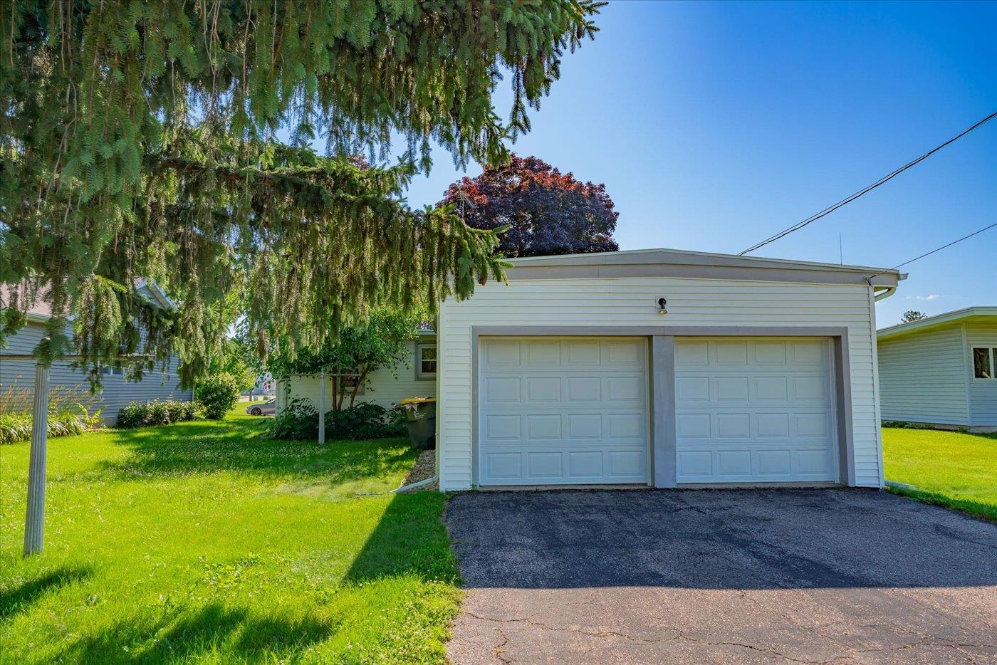 White detached two-car garage with paneled doors, asphalt driveway, and a large evergreen tree.