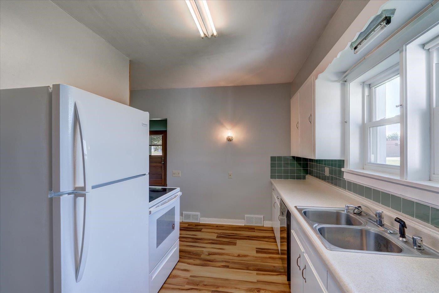 Kitchen with white appliances, green tile backsplash, and a double-basin sink beneath large windows.