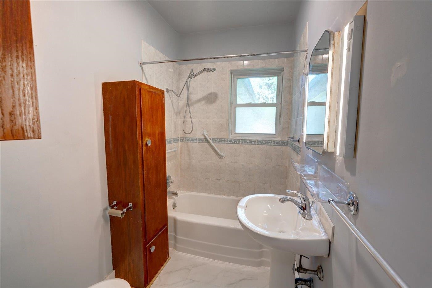 Bathroom with tiled tub and shower, pedestal sink, grab bars, and a tall wooden cabinet.