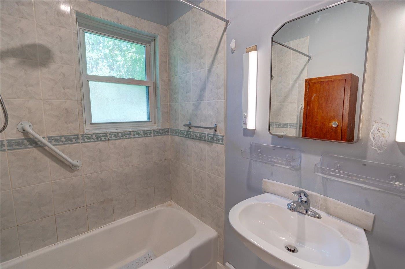 Bathroom with a tiled bathtub, frosted window, grab bars, and white sink beneath a large mirror.