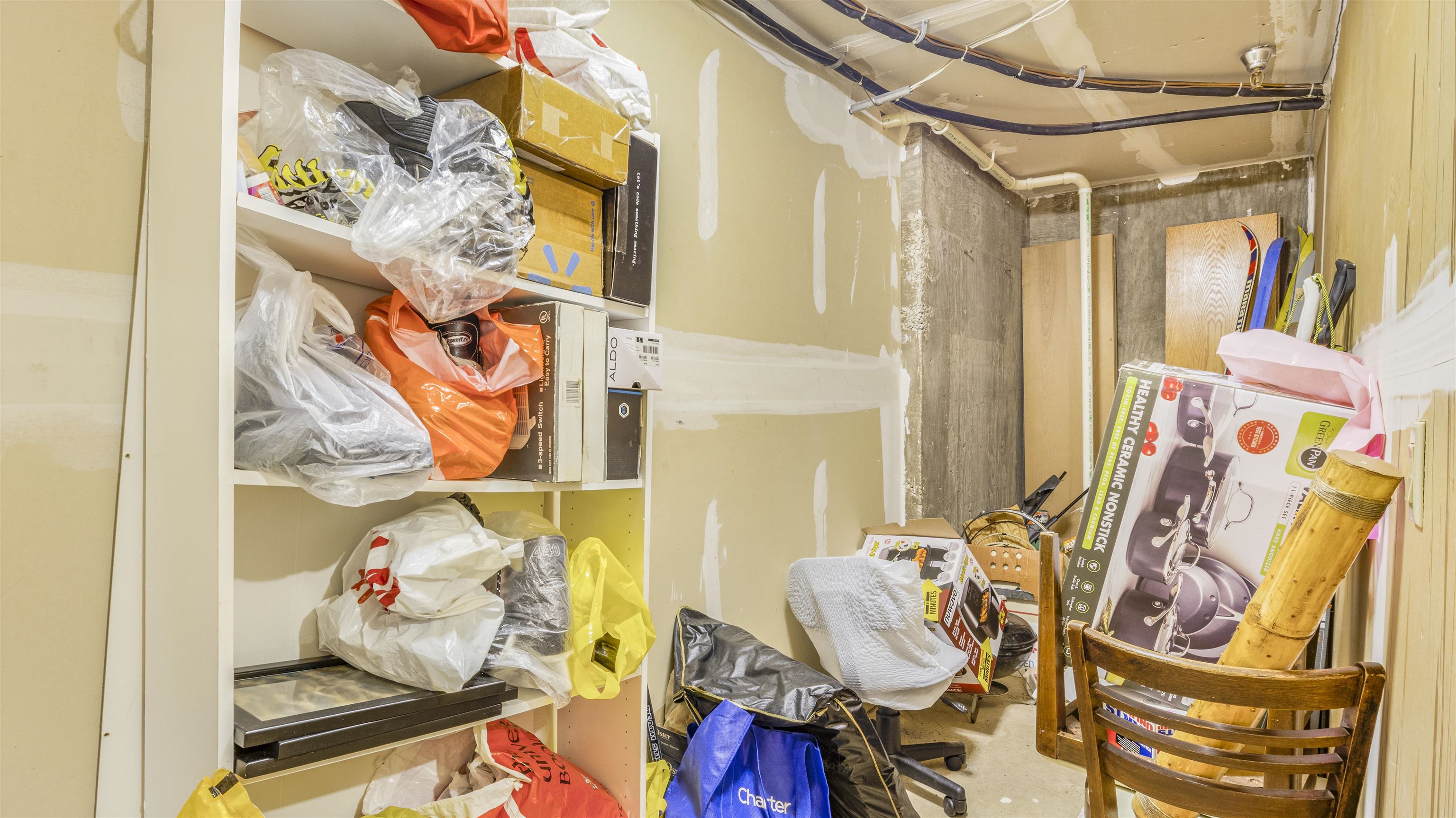 Unfinished basement storage area with exposed pipes, crowded shelves, and a wooden chair.