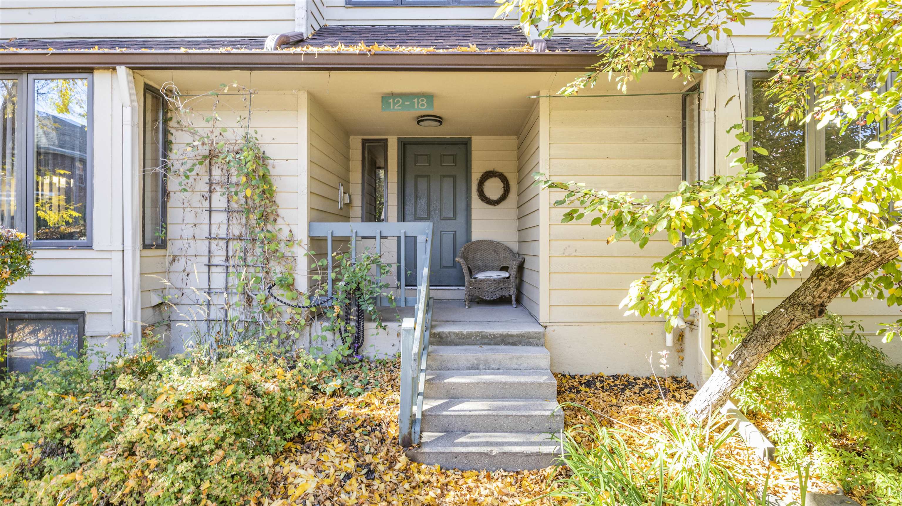 Home entrance with a covered porch, blue door, concrete steps, wicker chair, and autumn foliage.