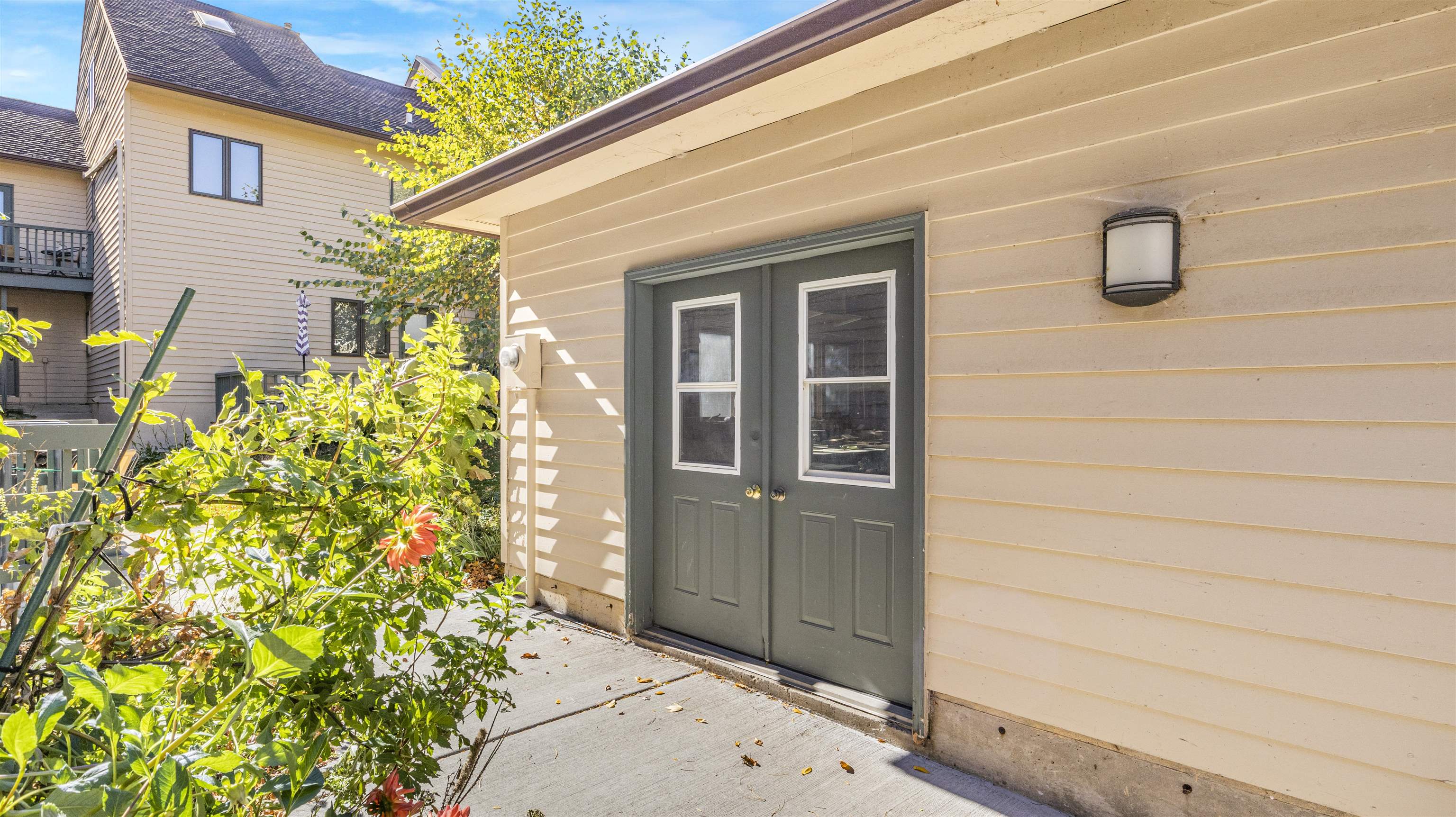 Beige-sided home with dark double doors opening onto a concrete patio beside a flowering garden.