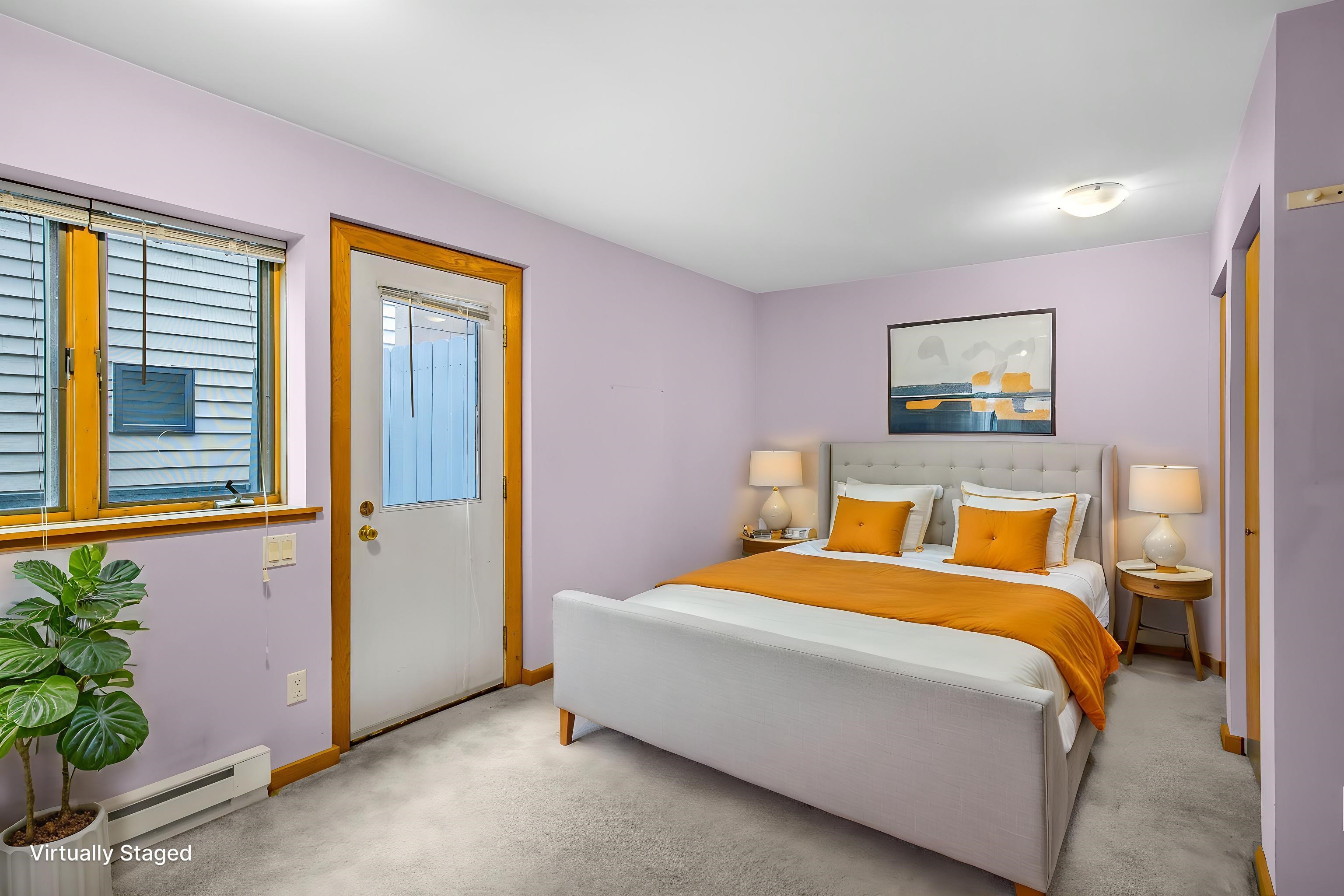 Bedroom with a white bed dressed in orange, lavender walls, and wood-trimmed windows.