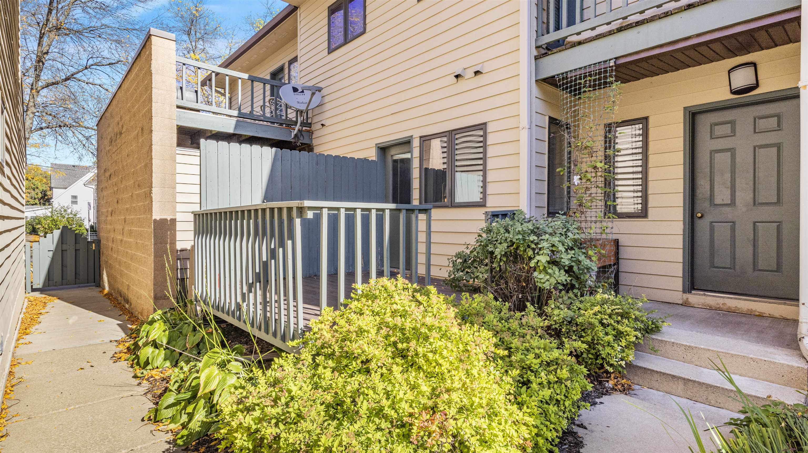 Residential building entry with gray doors, raised deck, concrete walkway, and landscaped shrubs.