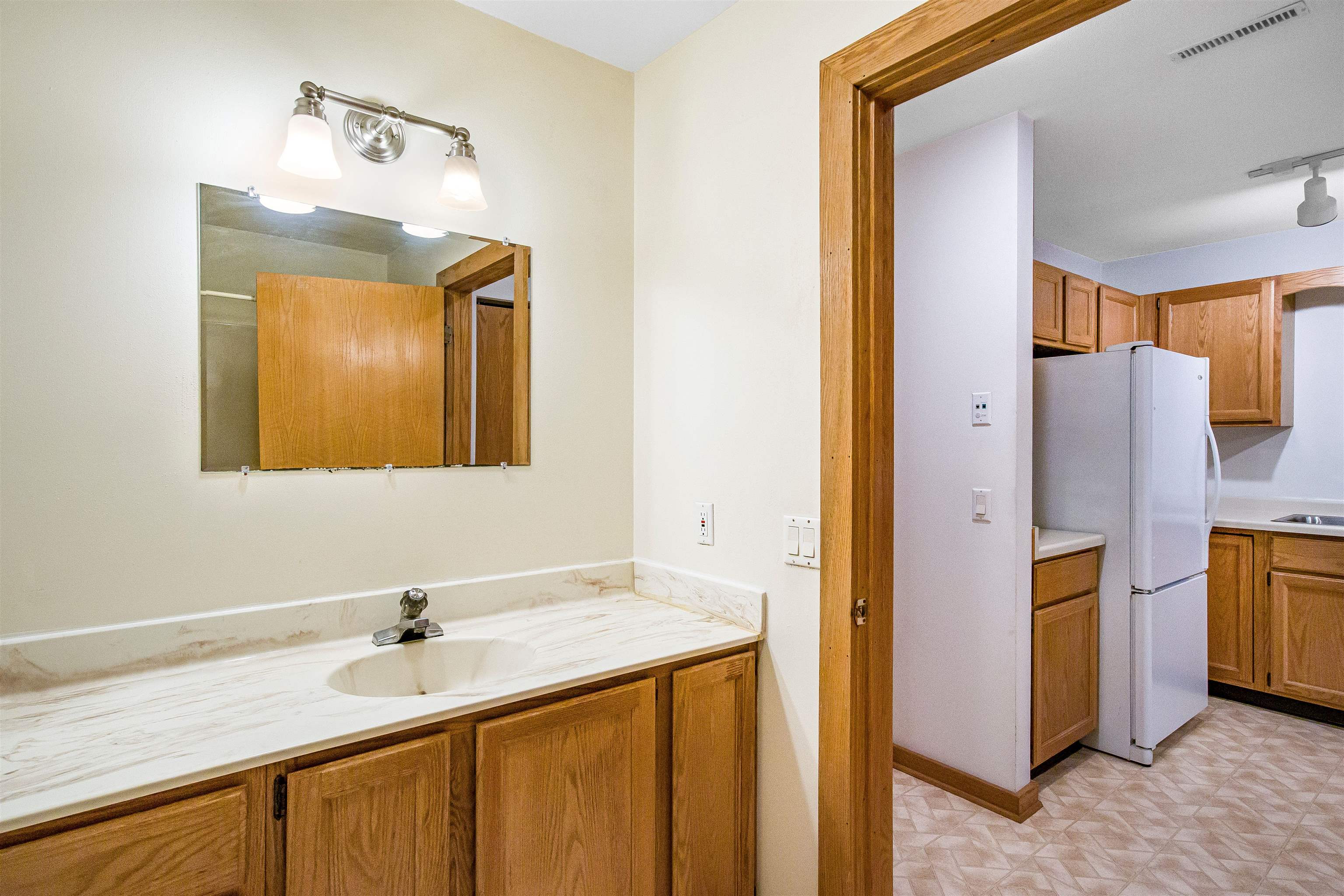 Bathroom vanity with a large mirror and wood cabinets, looking through to a kitchen with a white refrigerator.