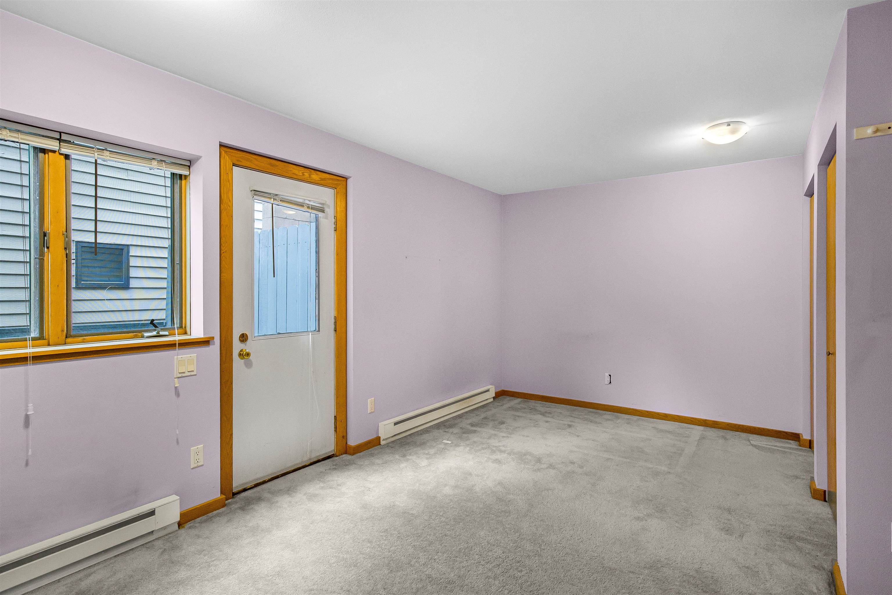 Empty carpeted room with lavender walls, wood-trimmed window and exterior door, and baseboard heaters.