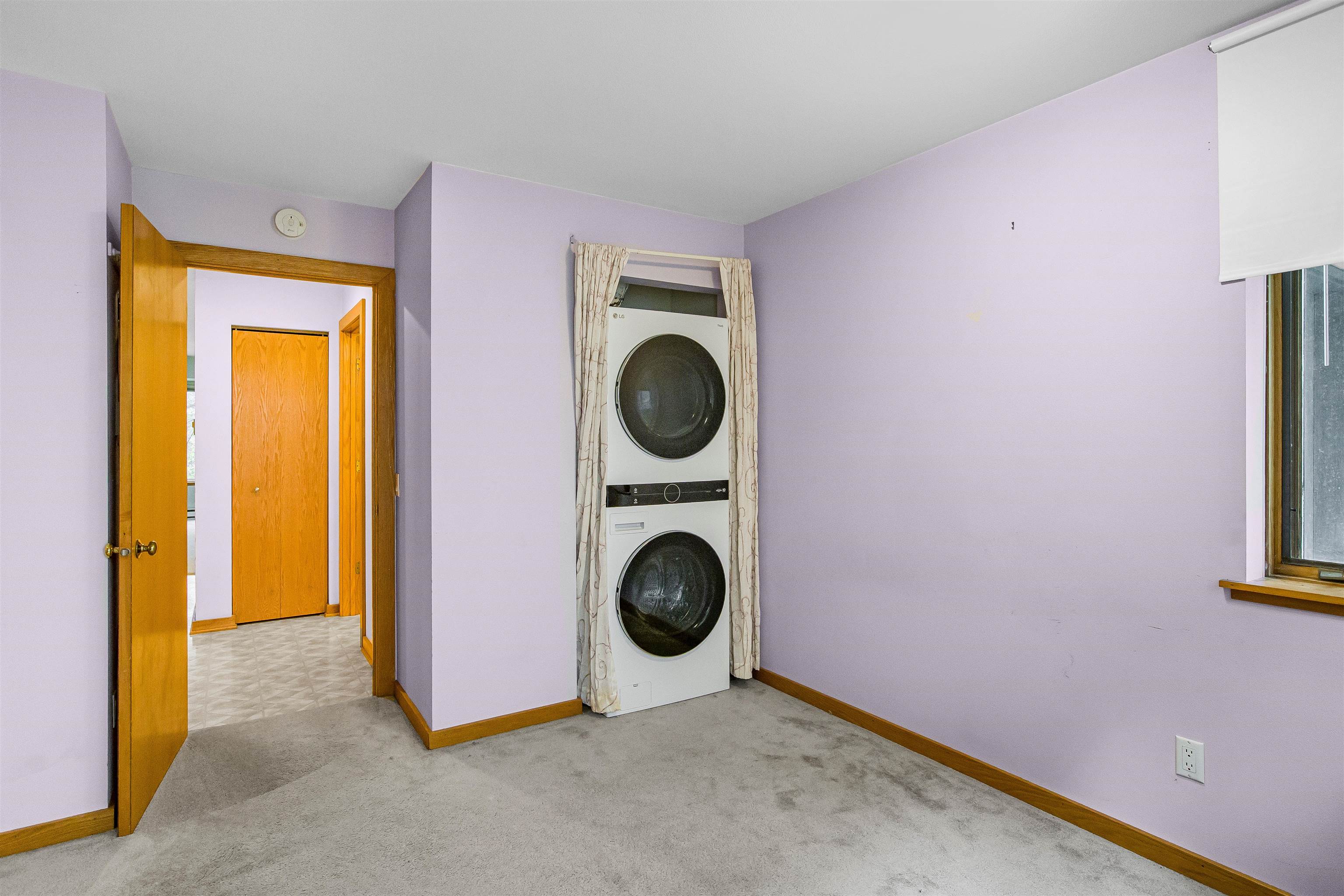 Carpeted room with a stacked washer and dryer behind curtains and a window with a lowered blind.