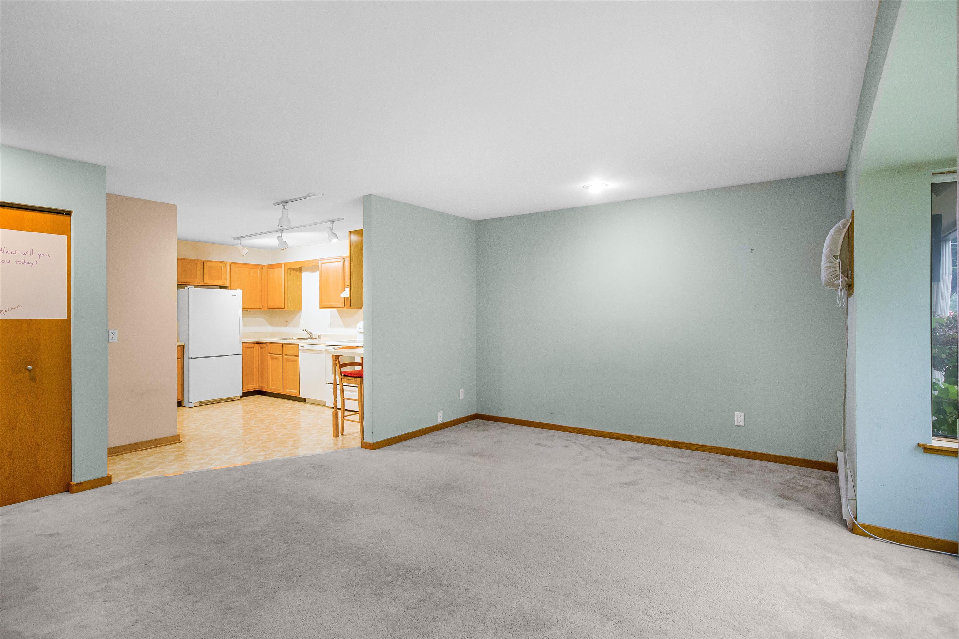 Empty carpeted living room opening onto a kitchen with honey-colored cabinets and a white refrigerator.
