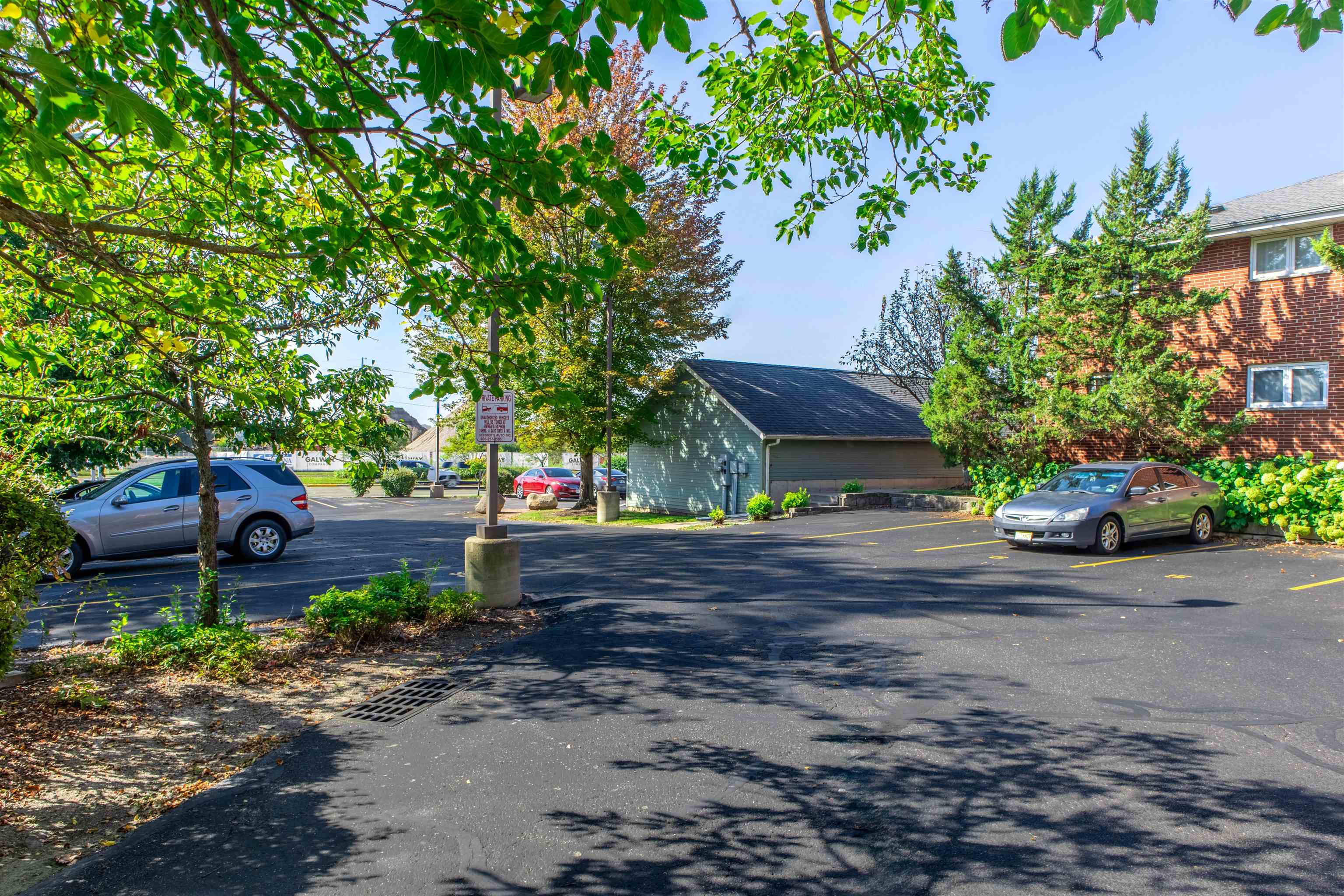 Parking area beside a brick residential building, with a detached garage, parked cars, and leafy trees.