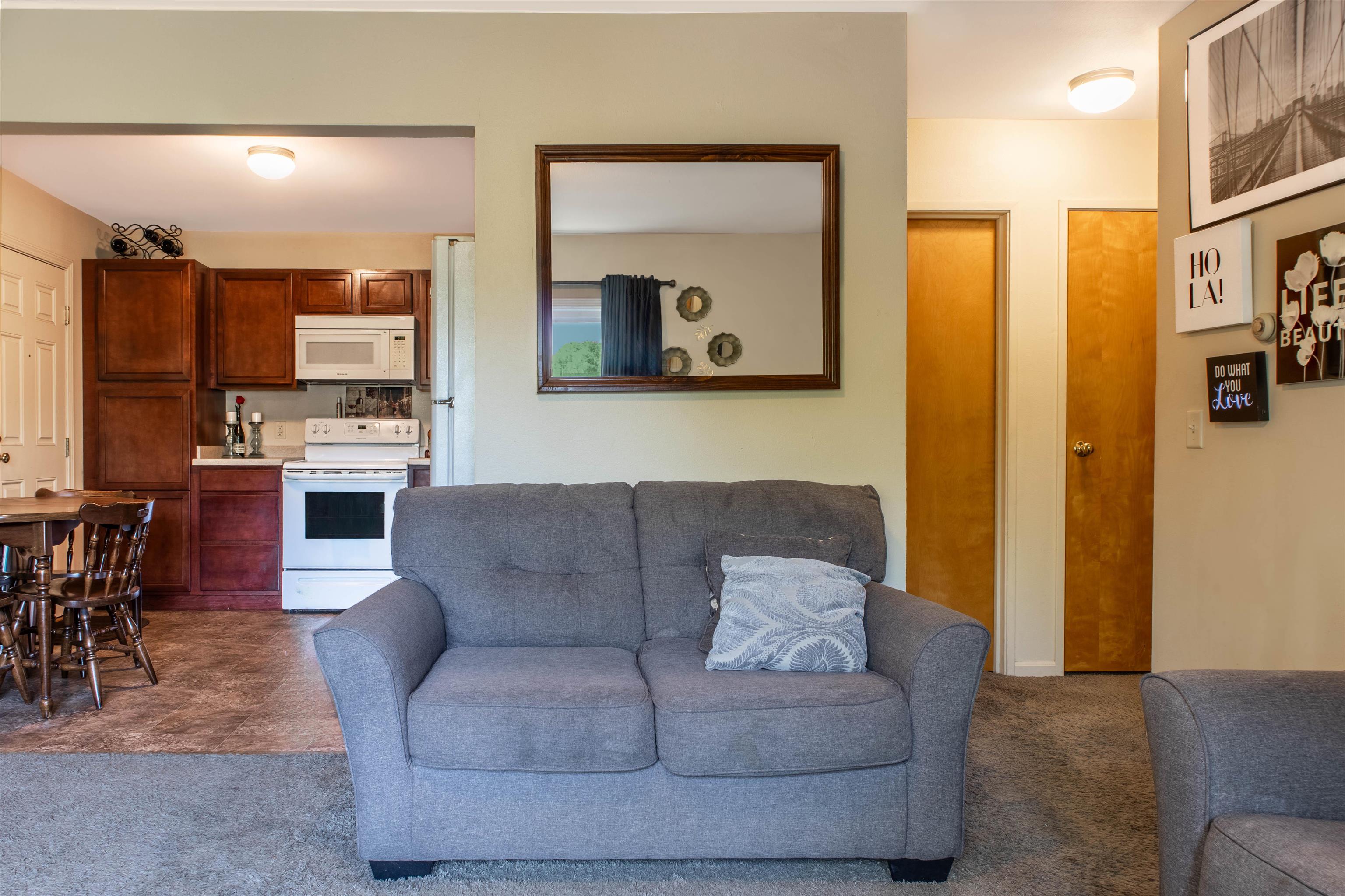 Furnished living room with gray loveseat, open kitchen, dark wood cabinets, and white appliances.