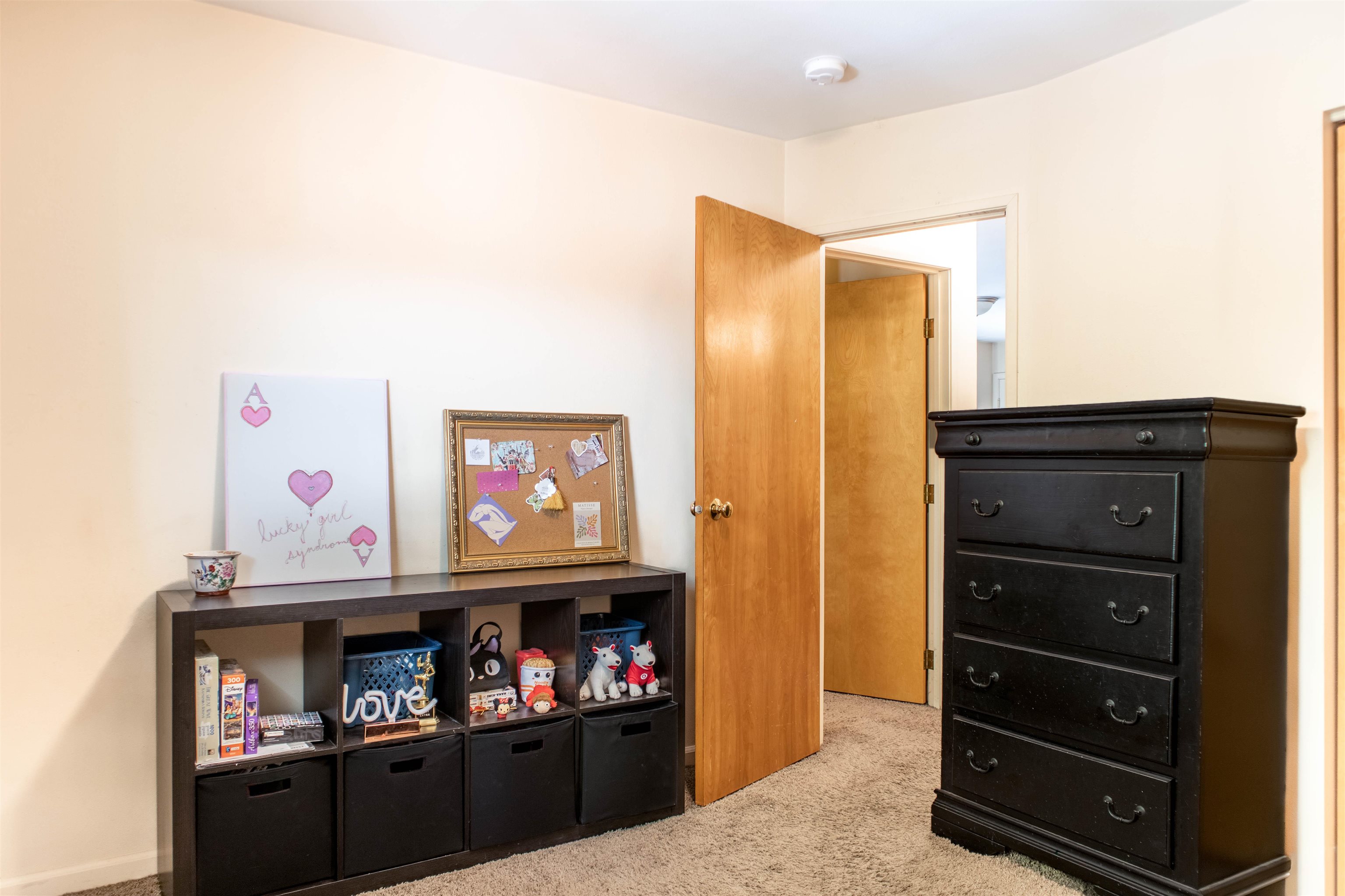 Bedroom with a black dresser, toy-filled cubby shelf, beige carpet, and open wooden door.