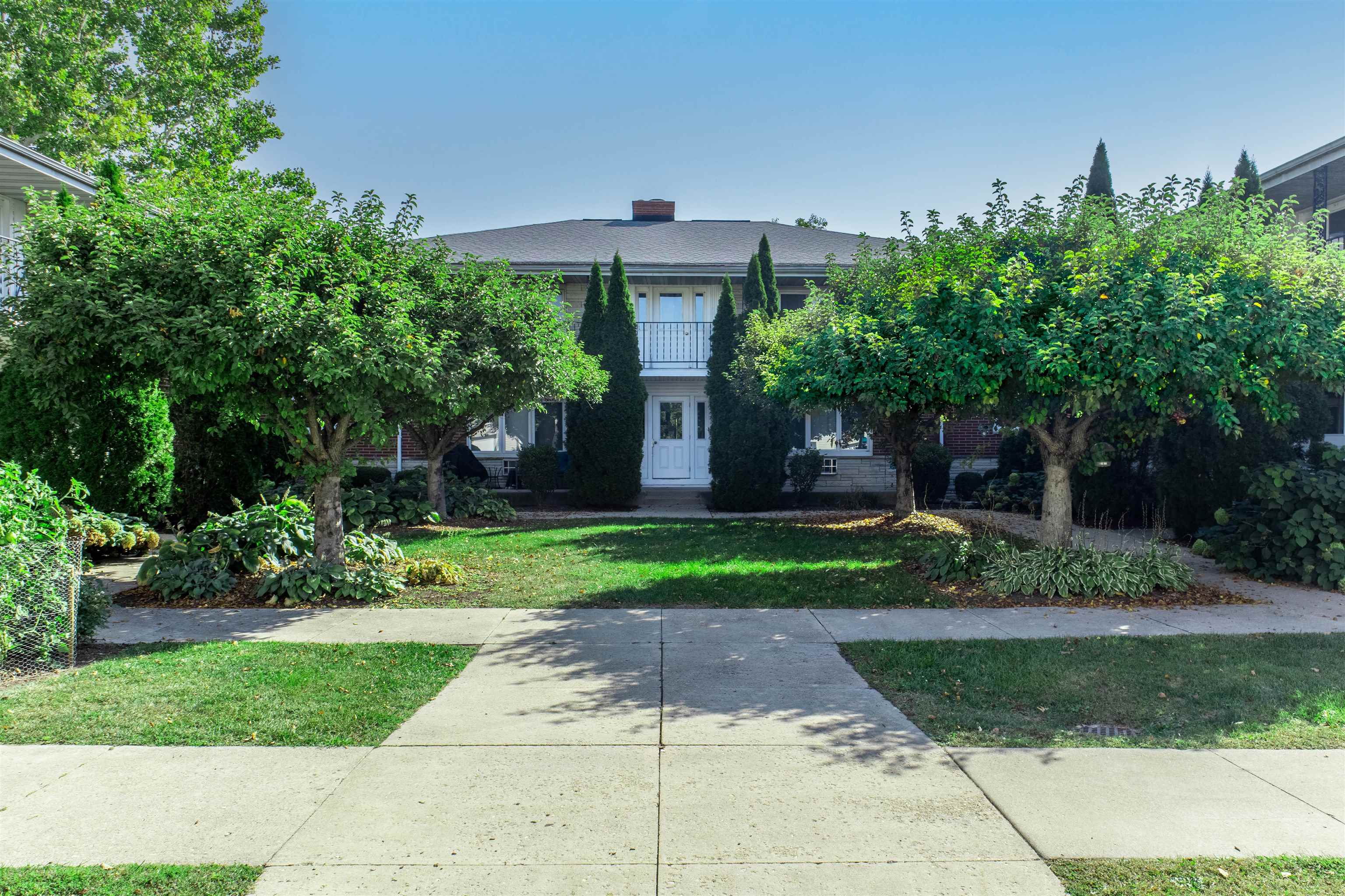 Home exterior with a central balcony and entry framed by mature trees and shrubs.
