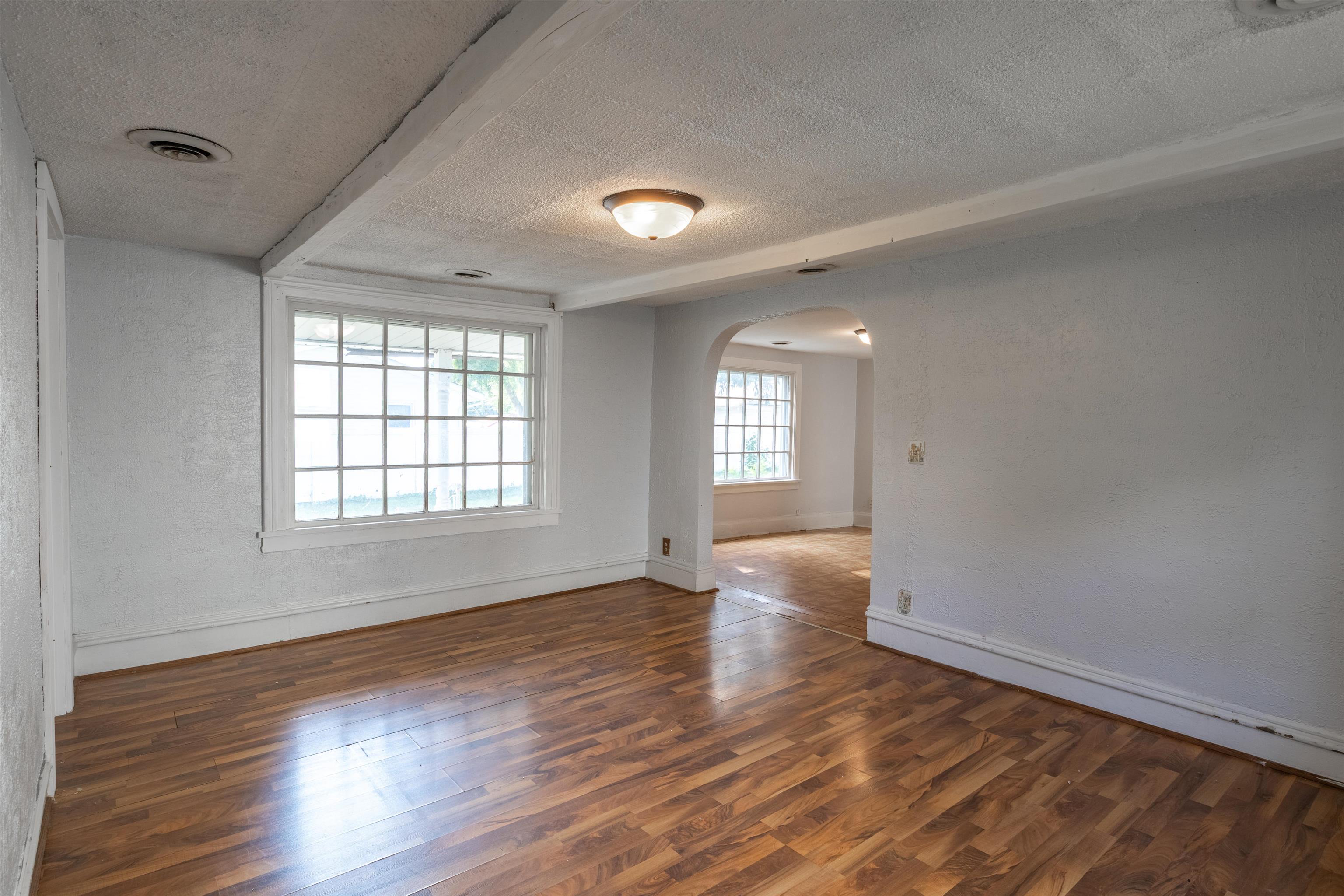 Empty living room with hardwood floors, divided-light windows, and an arched opening to another room.