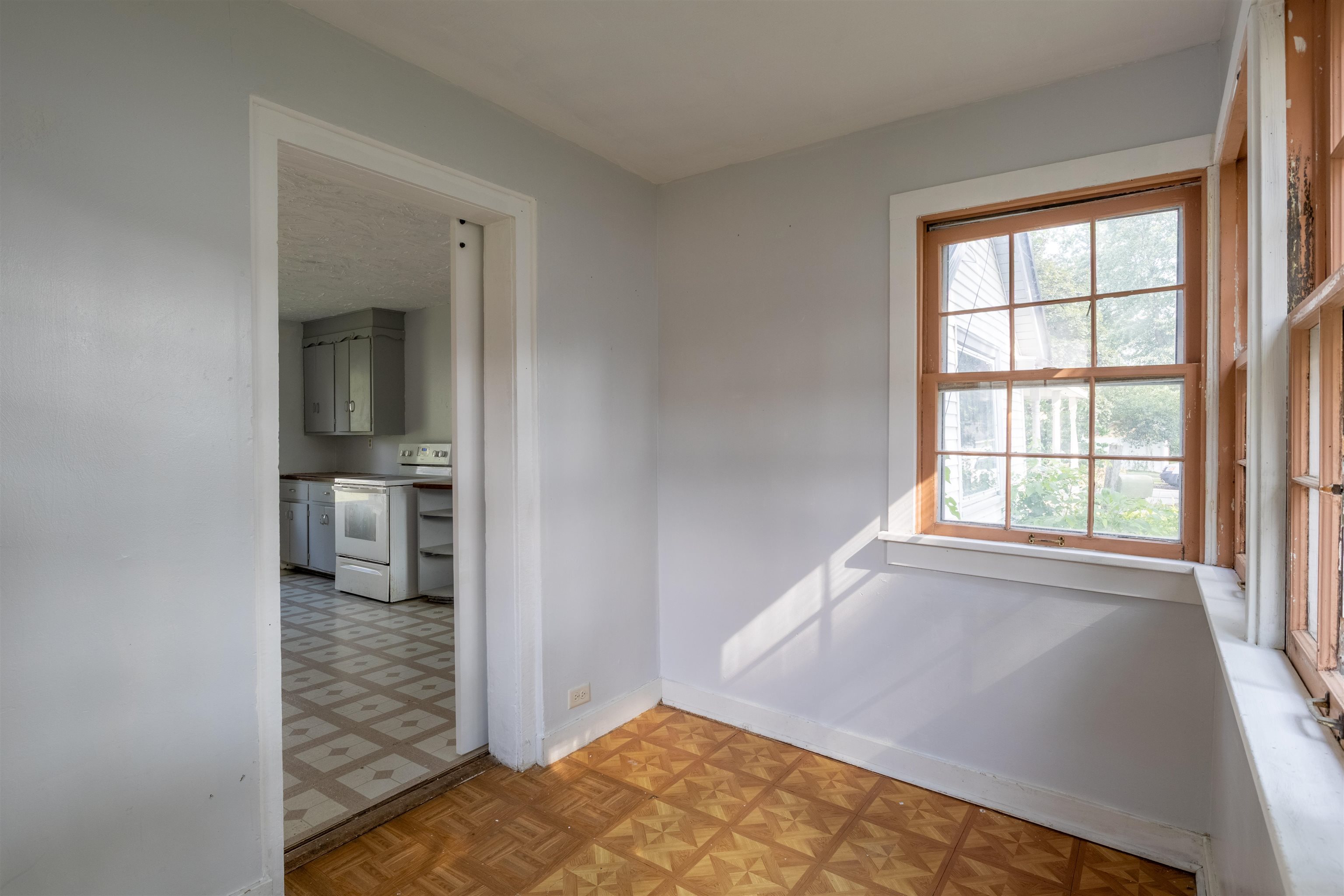 Empty room with patterned parquet flooring, a large wood-trimmed window, and a view into the kitchen.