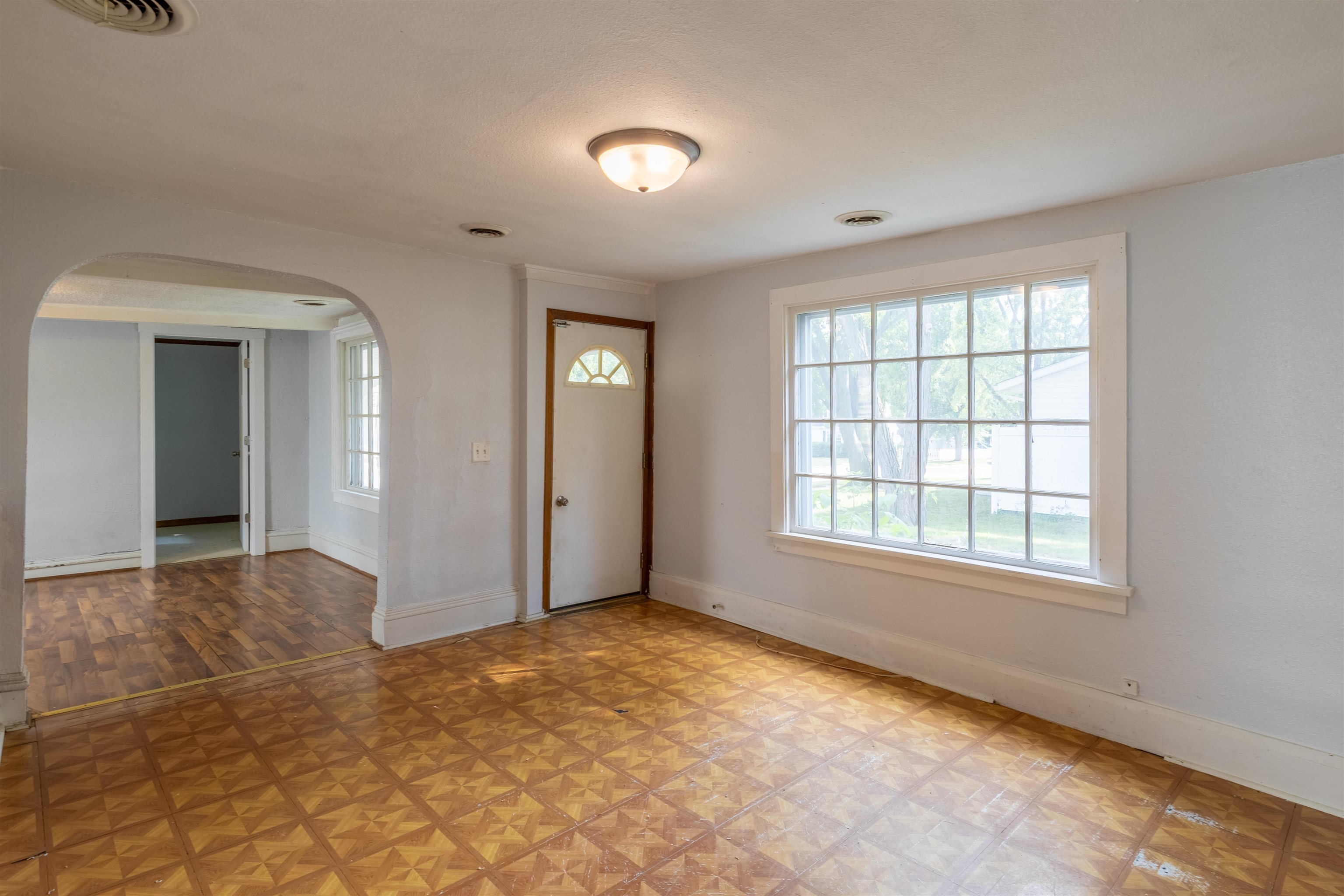 Empty room with patterned wood flooring, a large window, and an arched opening to an adjoining room.