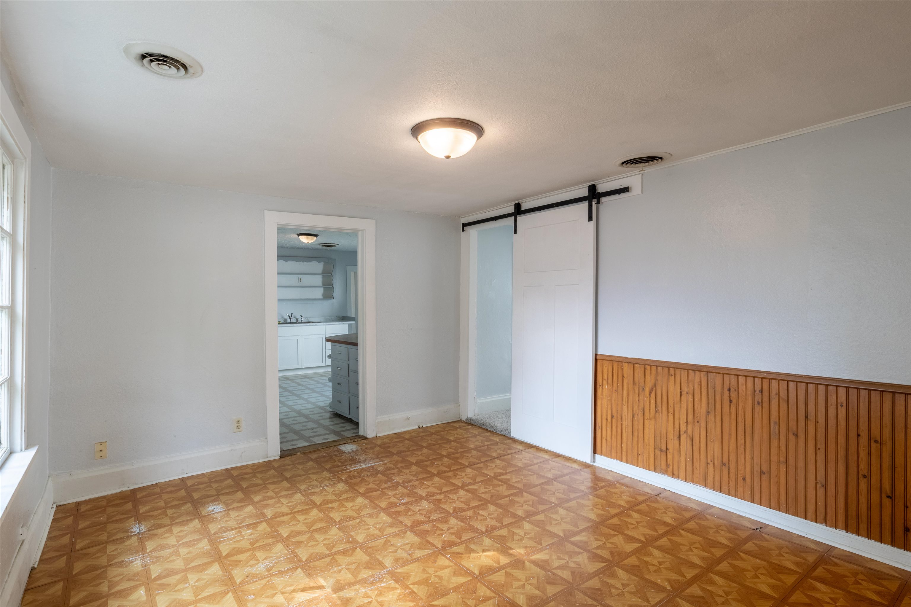 Empty room with parquet flooring, wood wainscoting, and a sliding barn door beside the kitchen doorway.