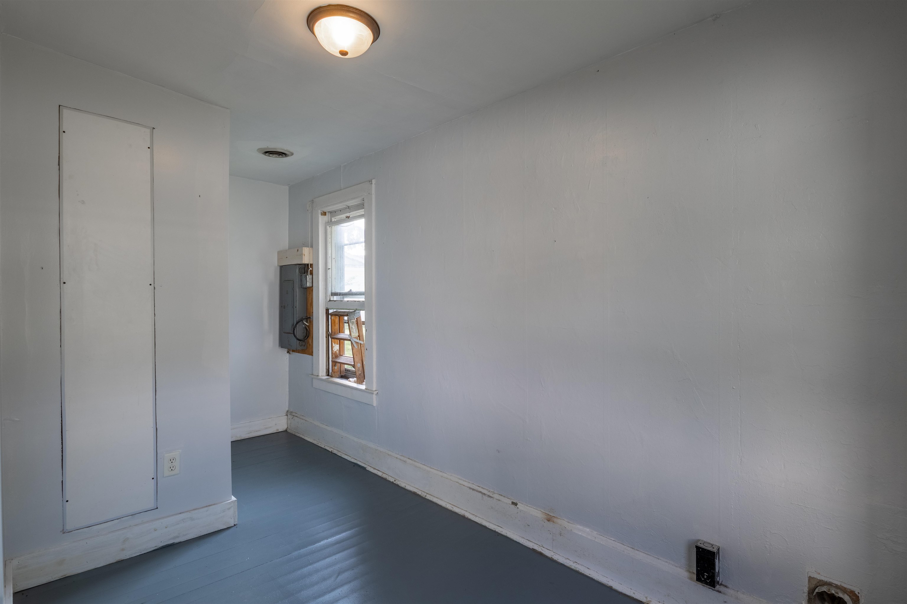 Empty narrow room with blue-gray floors, a small window, and an exposed electrical panel.