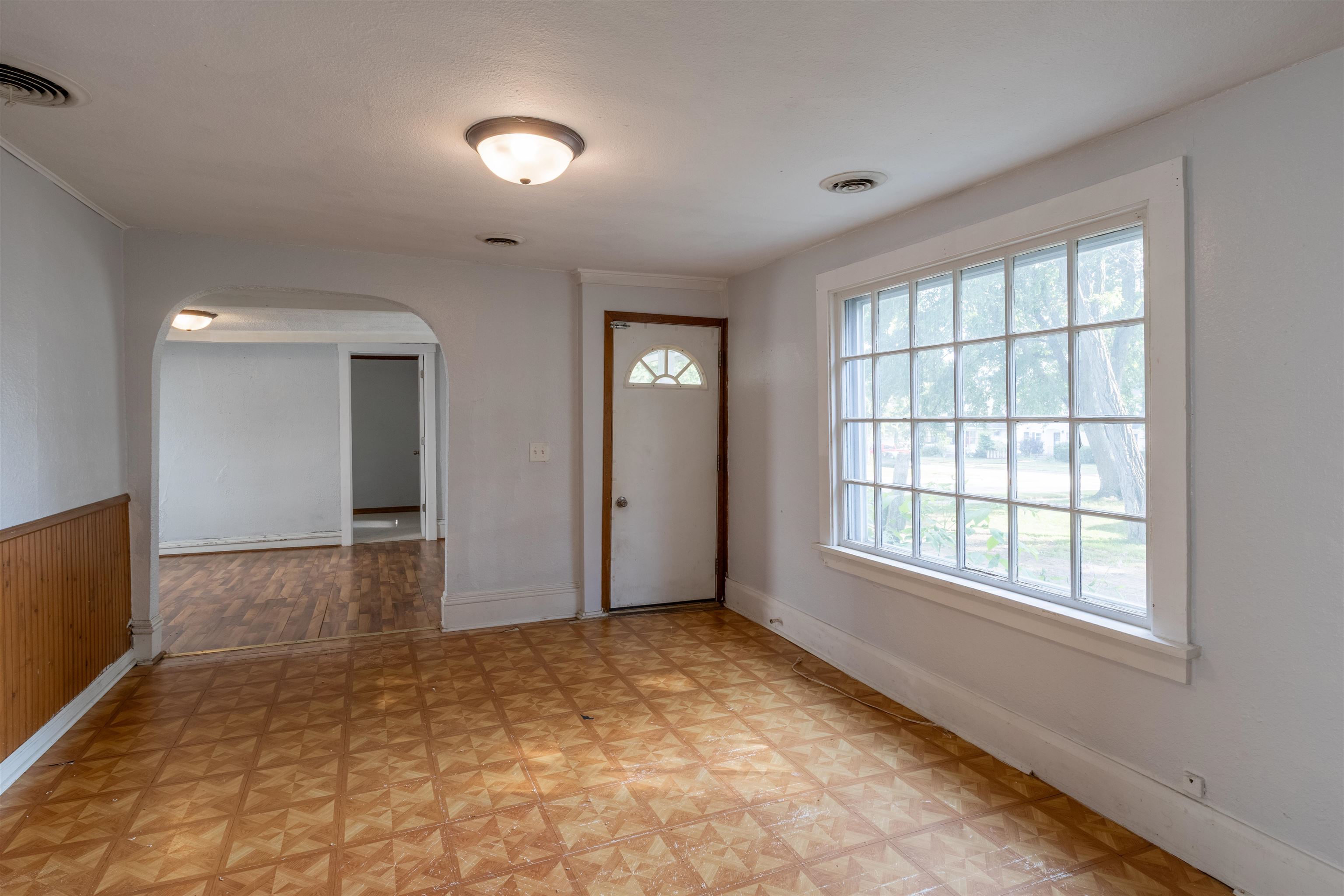 Empty front room with patterned flooring, wood wainscoting, a large window, and an arched opening.