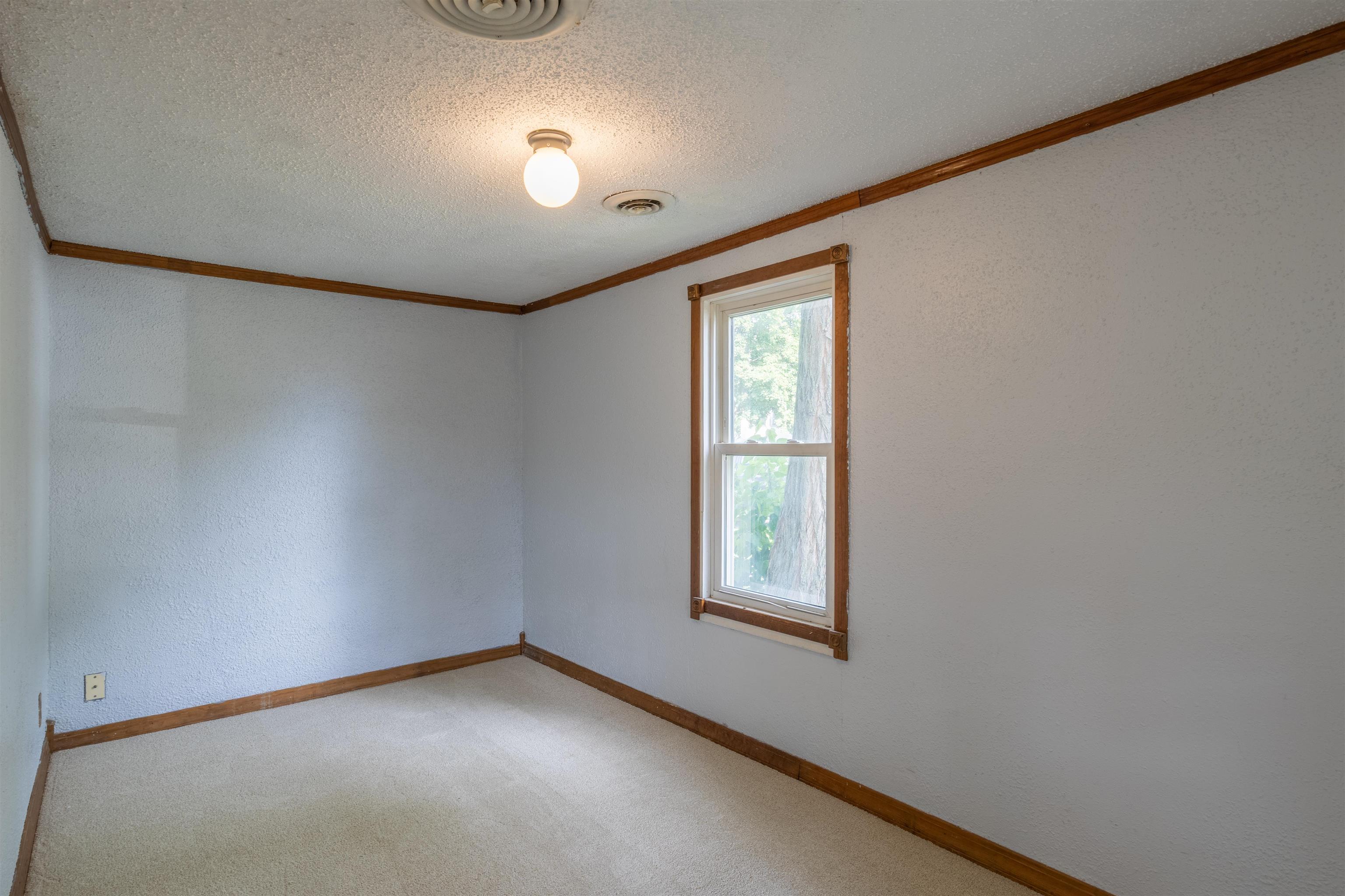 Empty carpeted room with dark wood trim and a window overlooking leafy trees.