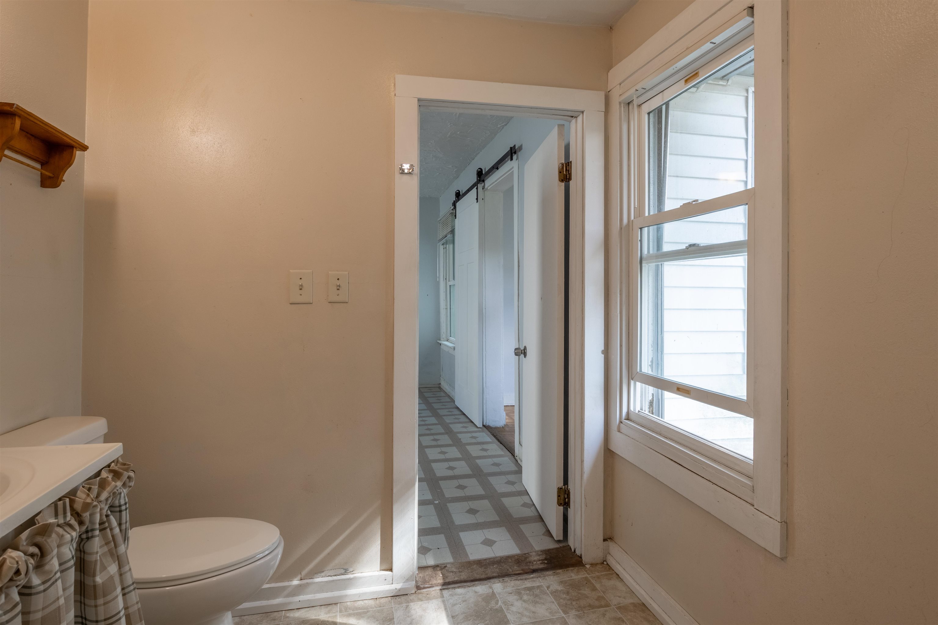Bathroom with a toilet, curtained sink, tall window, and doorway to a tiled hall.