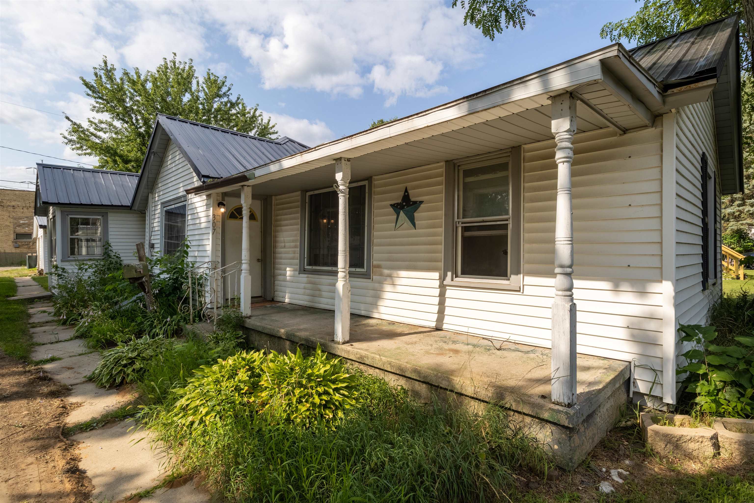 White-sided house with a covered porch, decorative columns, dark metal roof, and overgrown front garden.