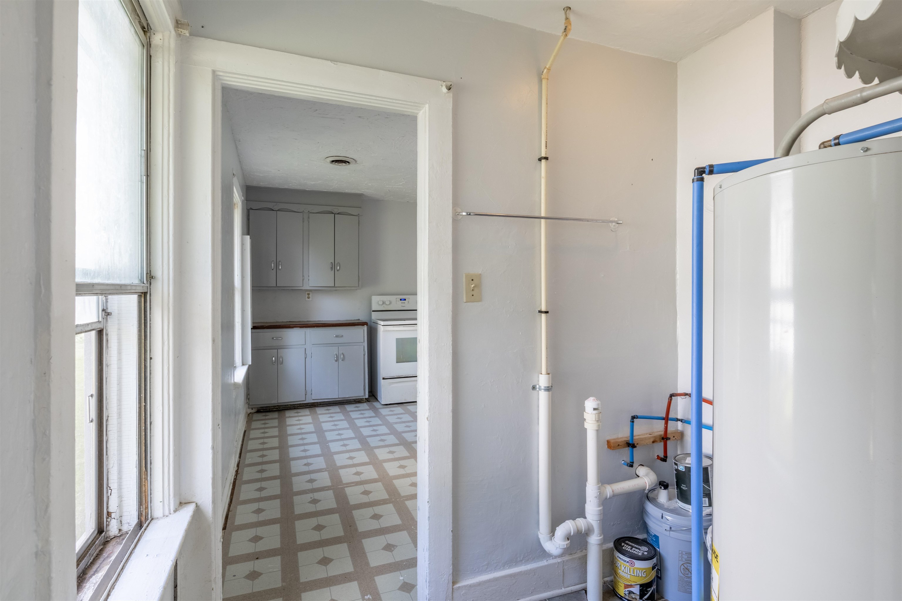 Utility room with a large water heater and exposed pipes beside a doorway to a gray-cabinet kitchen.