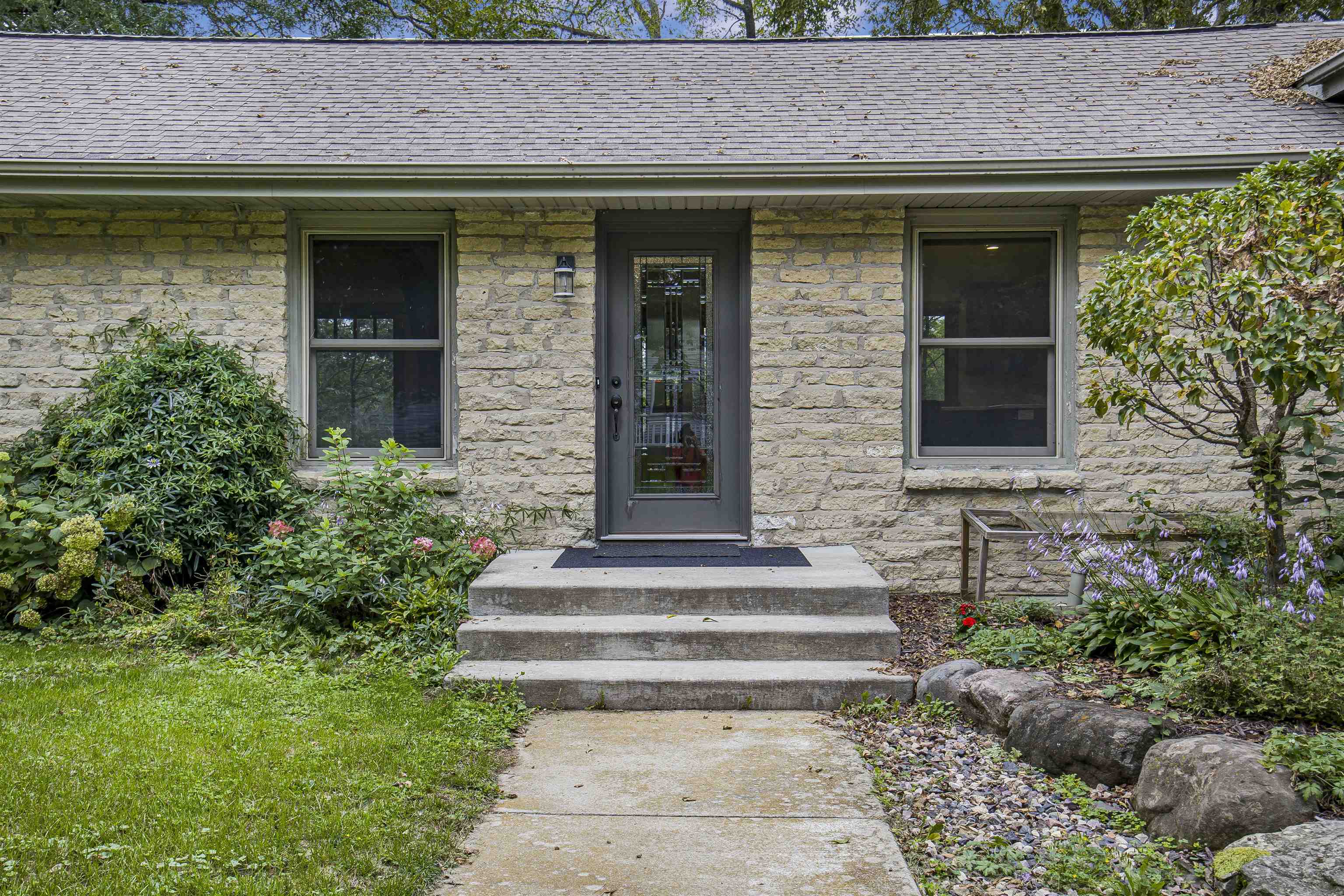 Stone house entrance with a dark glass-paneled door, concrete steps, and landscaped garden beds.