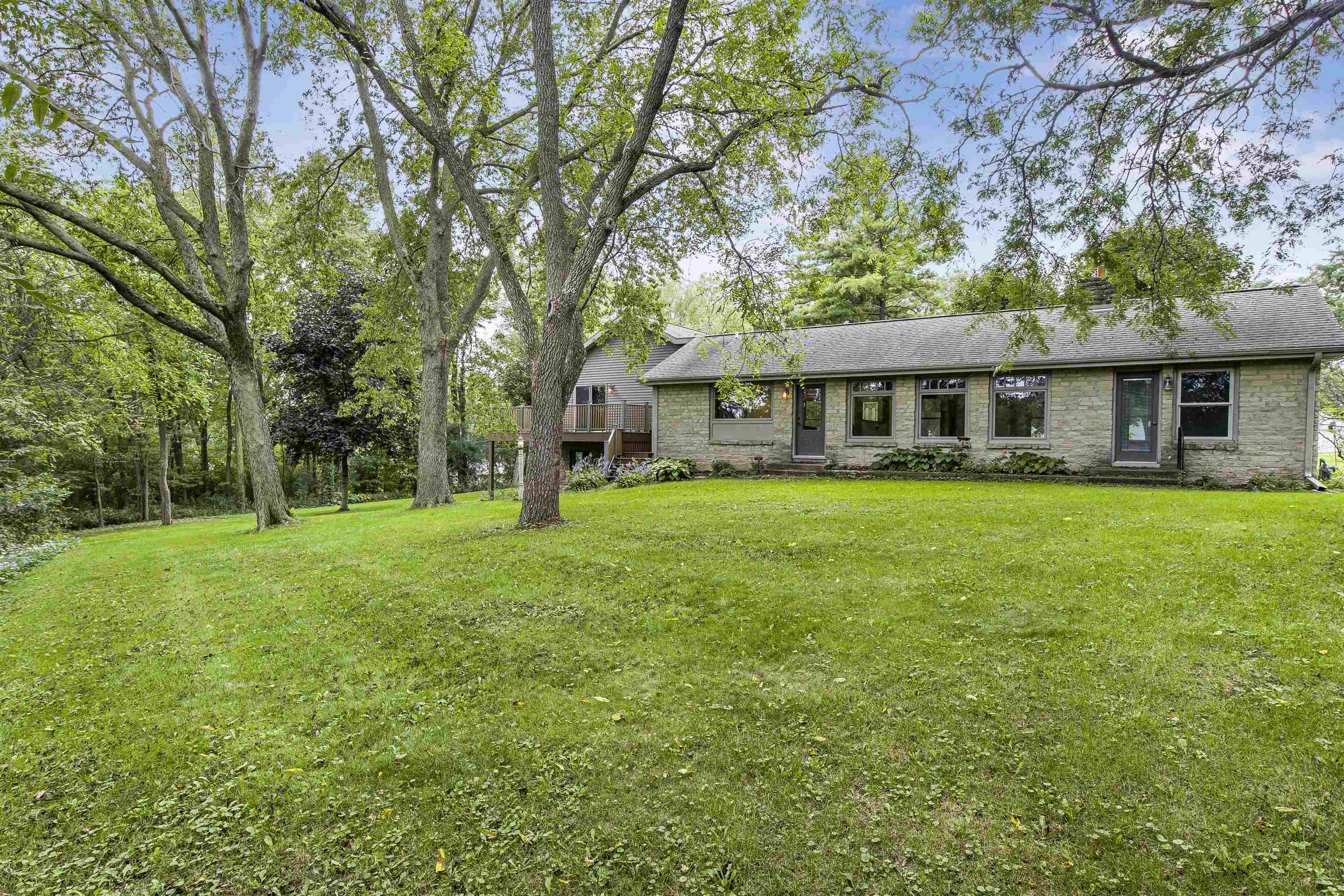 Single-story stone-sided home behind a broad lawn, framed by mature trees, with a raised wooden deck.