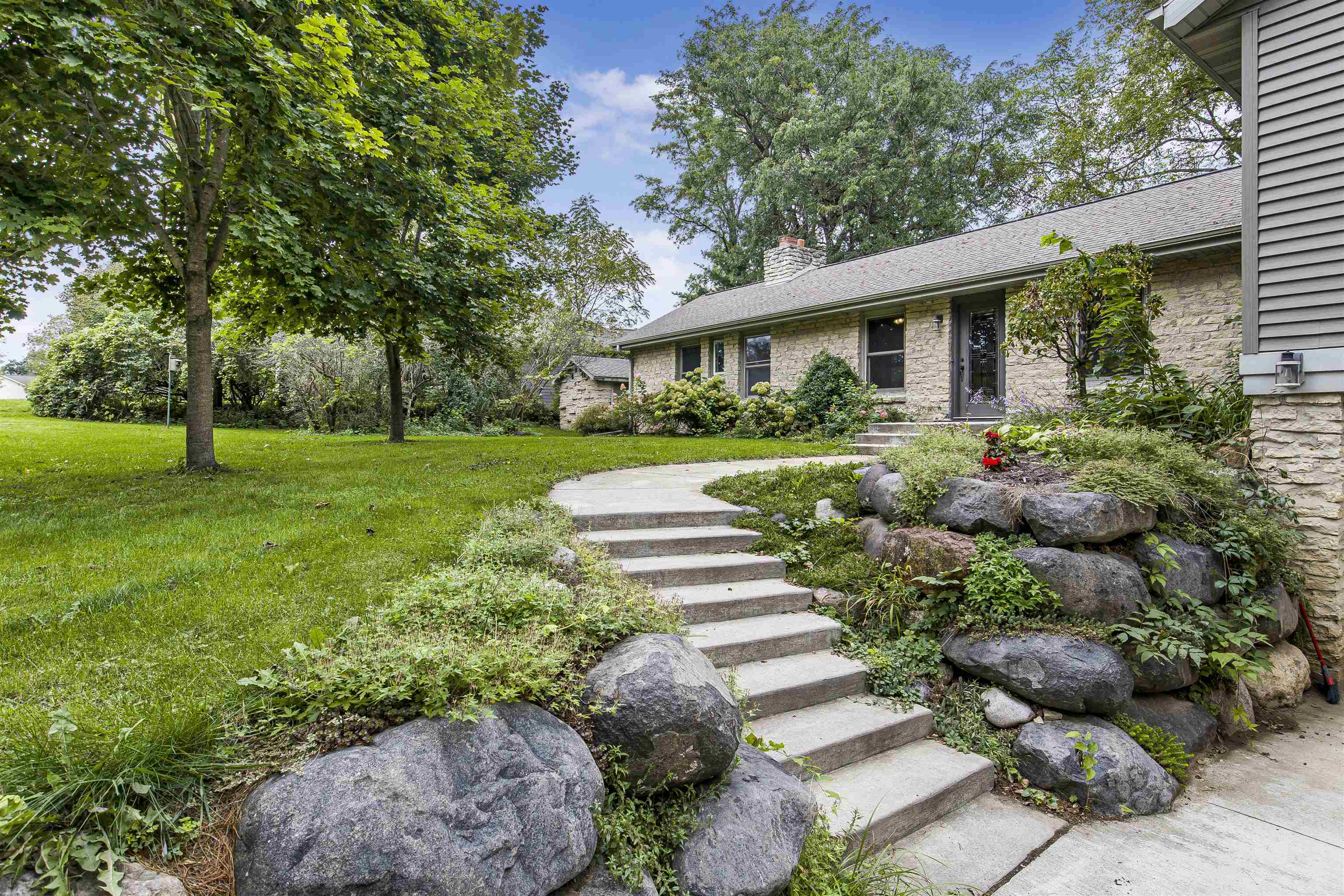 Stone house entrance with concrete steps winding through landscaped rocks and shrubs beside a leafy lawn.
